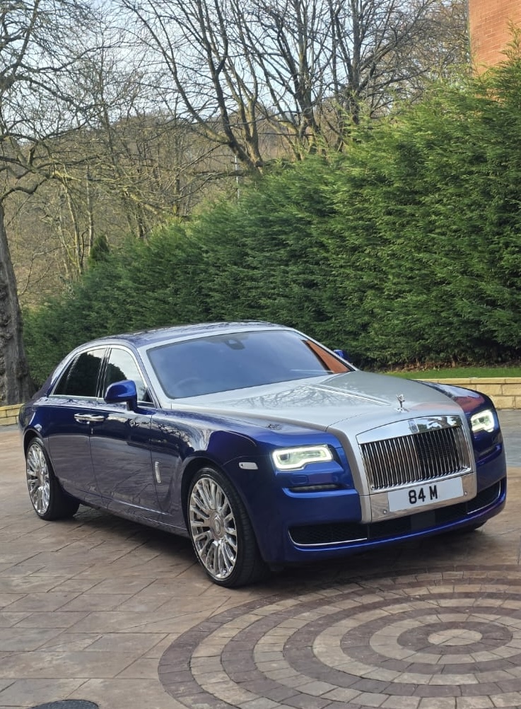 A luxurious blue and silver Rolls-Royce with shiny rims parked on a patterned stone driveway, framed by lush greenery and soft daylight.
