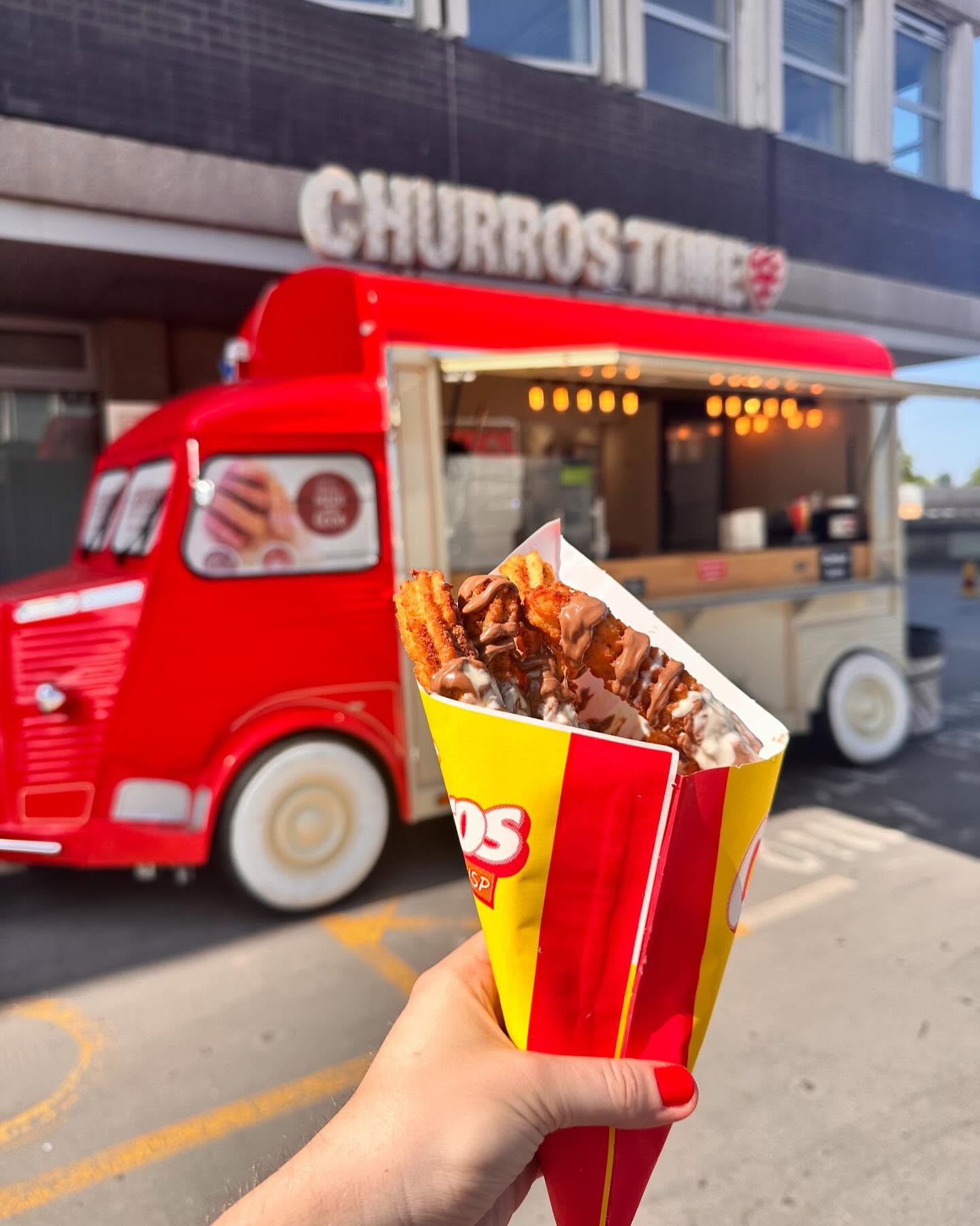 Hand holding chocolate-drizzled churros in a red-yellow paper cone before a bright Churros Time vintage van catering truck, warm daylight.