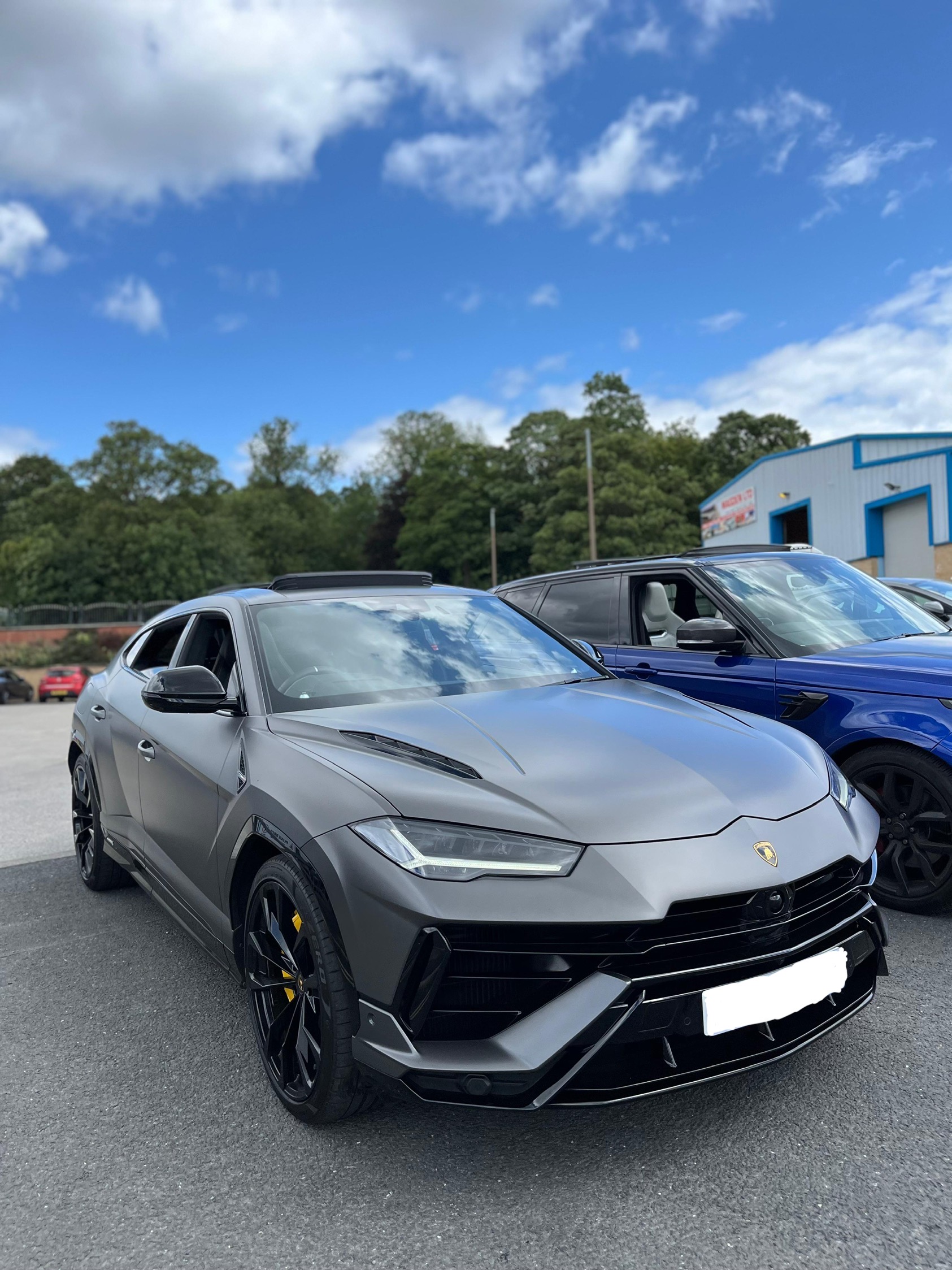 A sleek, matte gray Lamborghini Urus gleams under a bright blue sky, showcasing its powerful lines and luxury design amidst a casual gathering.