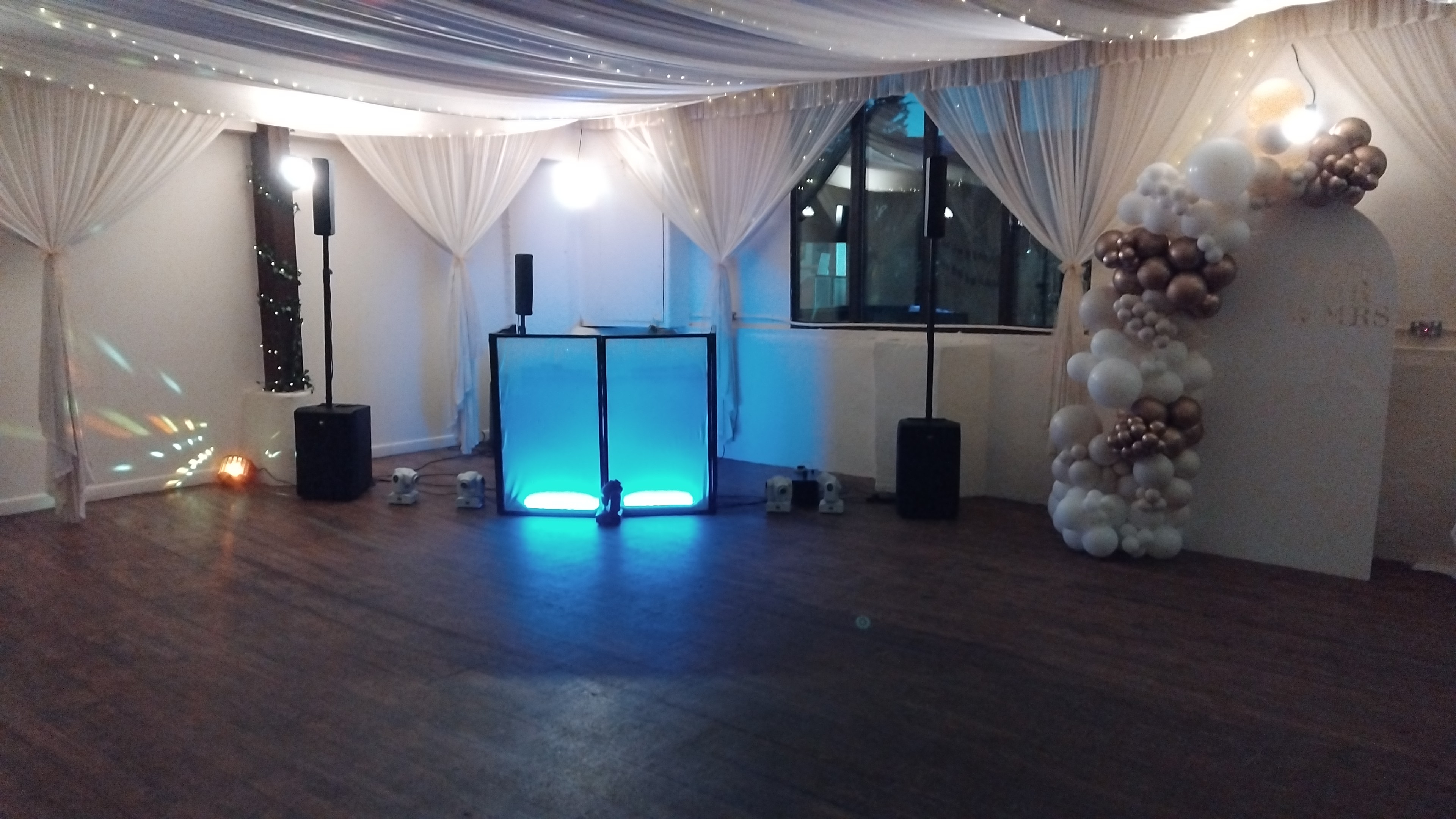A softly lit venue features a sleek DJ booth with vibrant blue accents, flanked by speakers and elegant balloon decor, exuding charm.