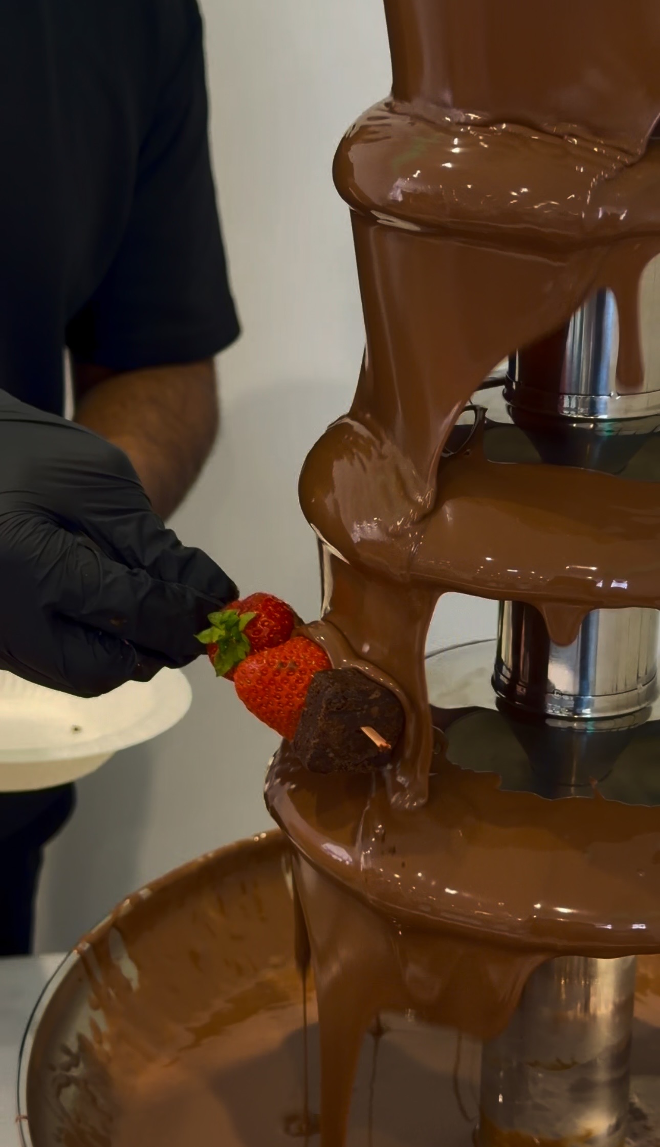 Elegant catering close-up of a chocolate fountain cascading over polished steel tiers, with gloved hands dipping strawberries in warm, soft light.