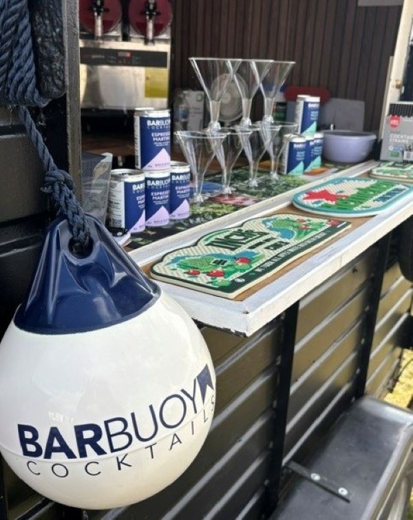 BarBuoys cocktail catering station with martini glasses, branded cans, and nautical trays on a white counter, under warm indoor lighting.