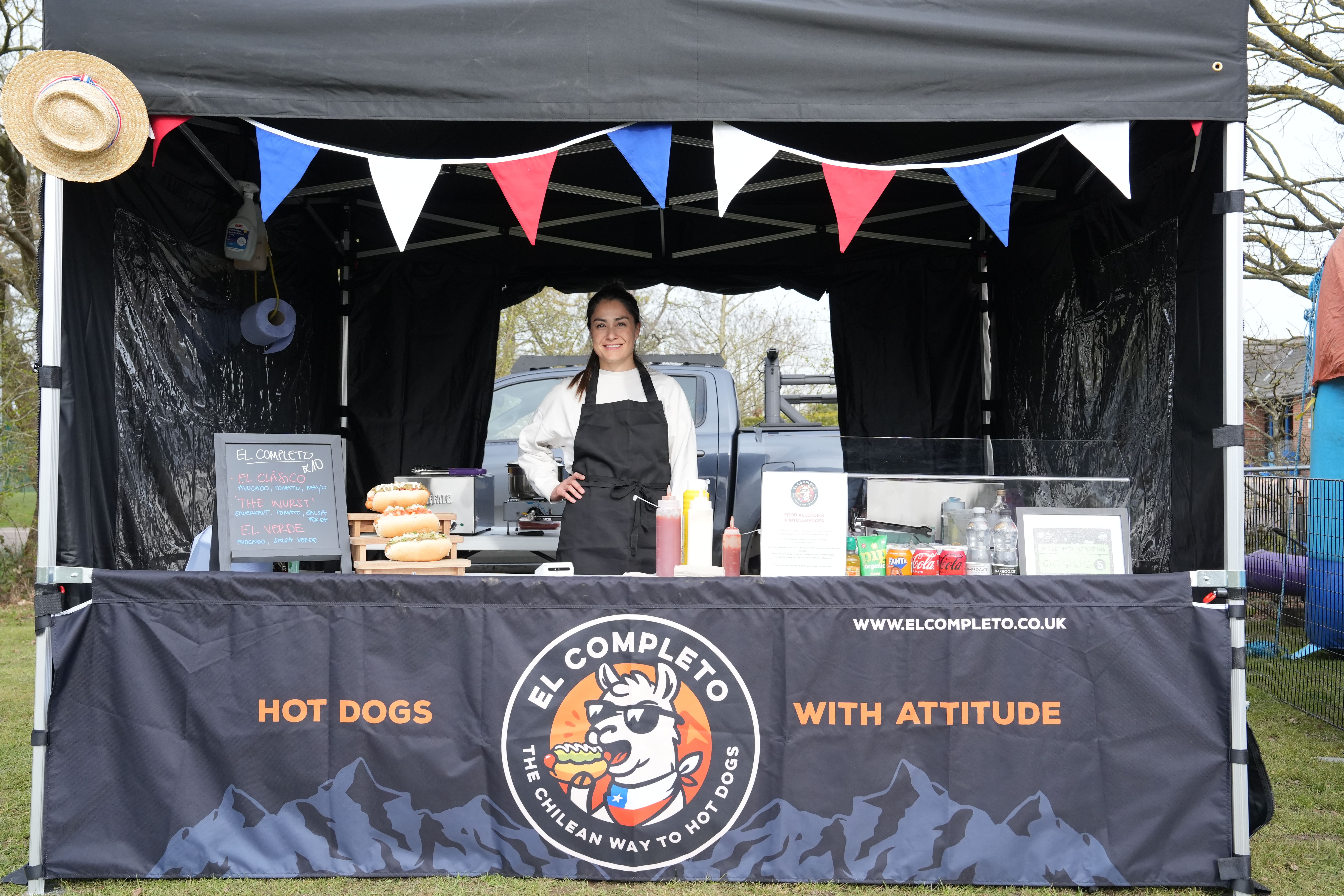 A vibrant food stall features neatly arranged hot dogs, colorful condiments in bottles, and a friendly vendor smiling under festive bunting.