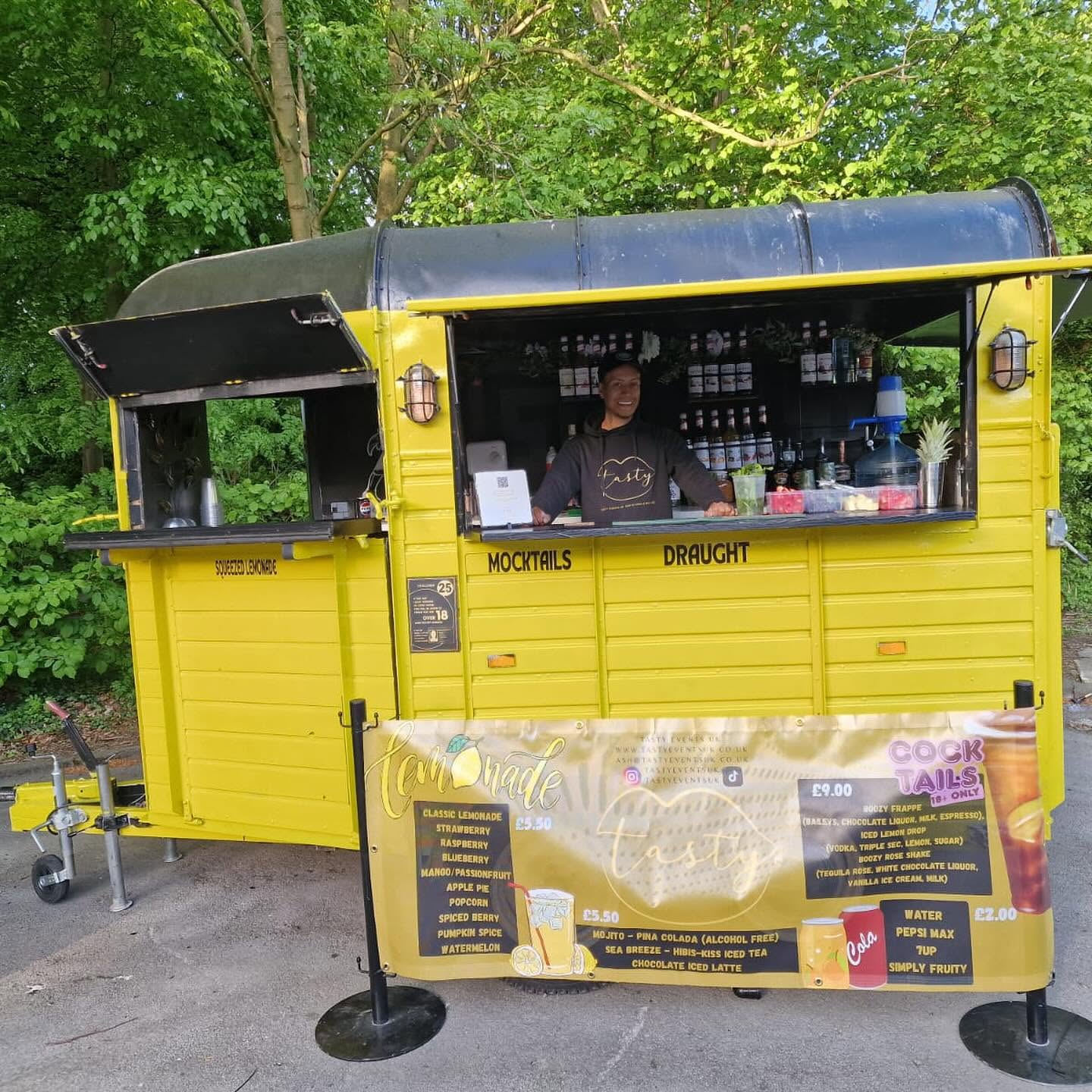 Bright yellow vintage drinks van serving lemonade, mocktails and draught cocktails, with polished jars and menus, set in a leafy park.