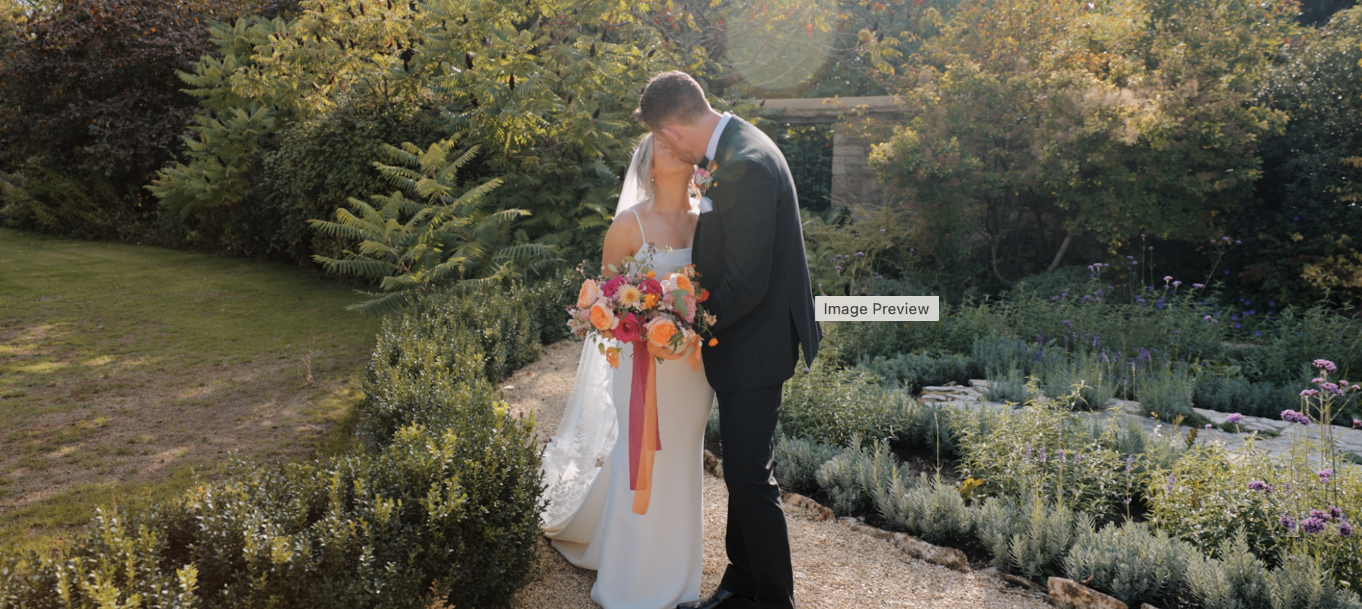 Sunlit garden wedding kiss with bride and groom, colorful bouquet, white gown, black suit, and lush greenery in a romantic outdoor setting.