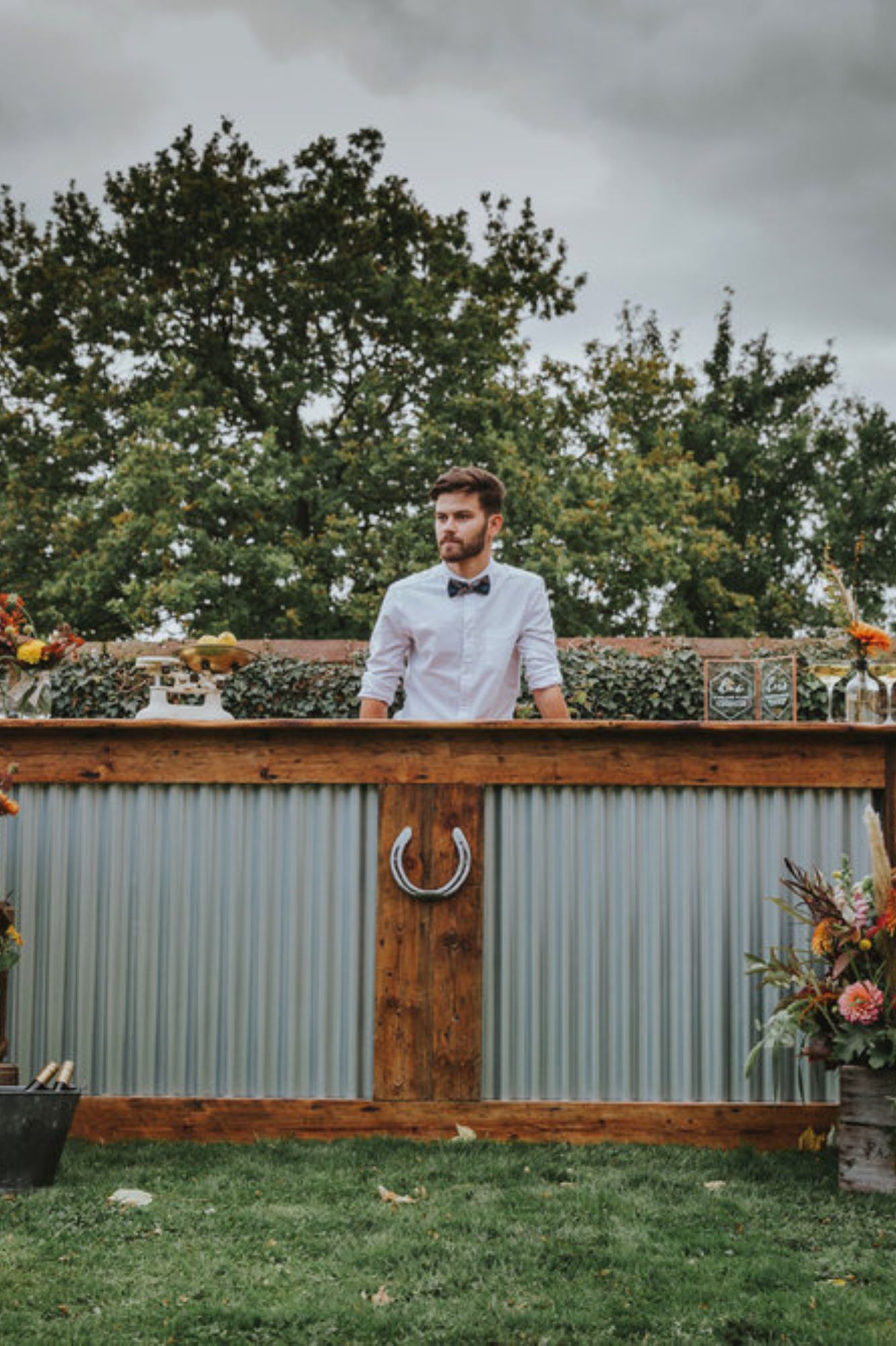 A rustic wooden bar with galvanized metal features a neatly dressed bartender. Fresh floral arrangements add color, enhancing the inviting atmosphere.