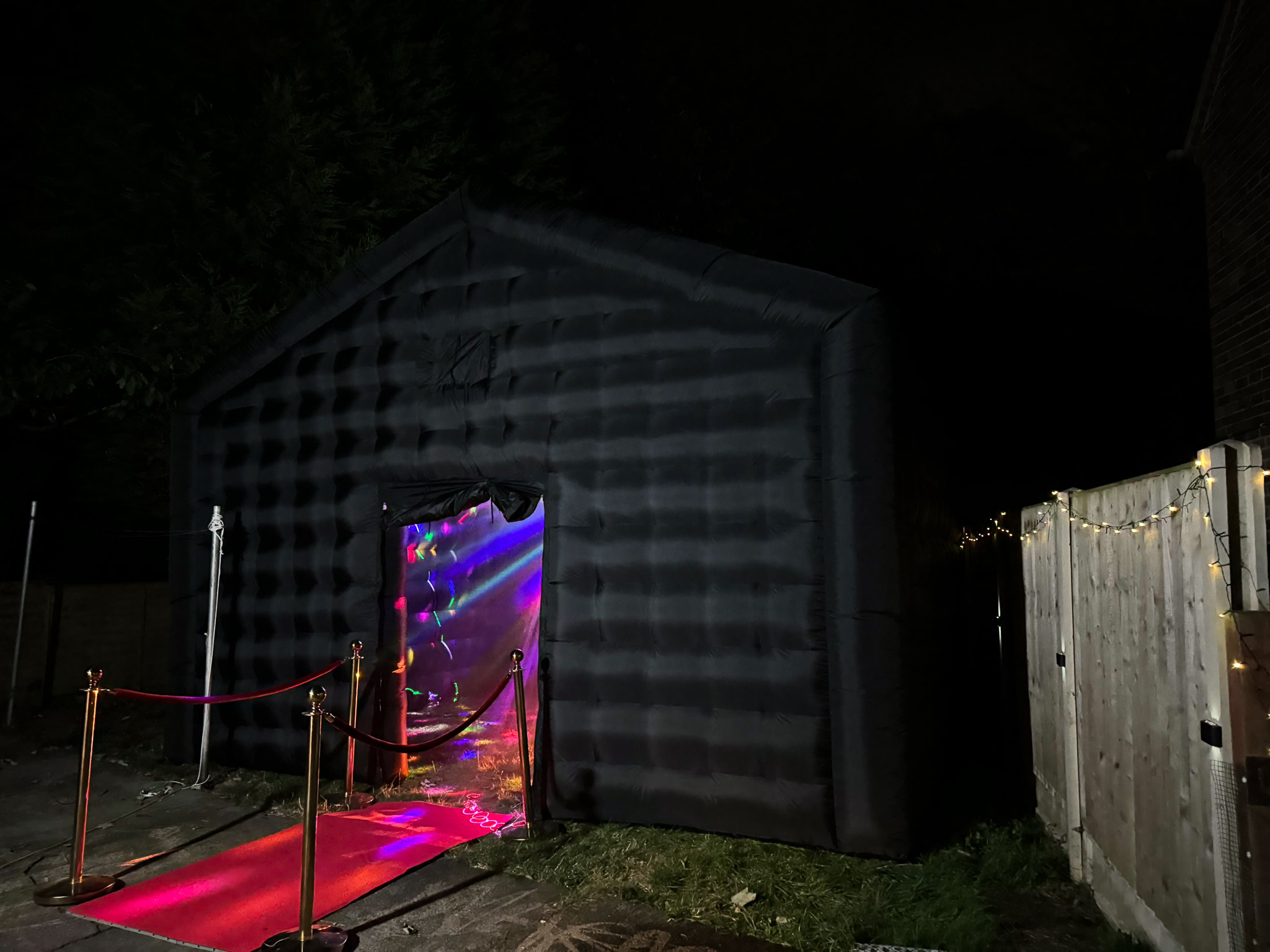 A dark inflatable structure features a vibrant entrance with colorful lights, flanked by a red carpet and golden stanchions, set for a festive event.