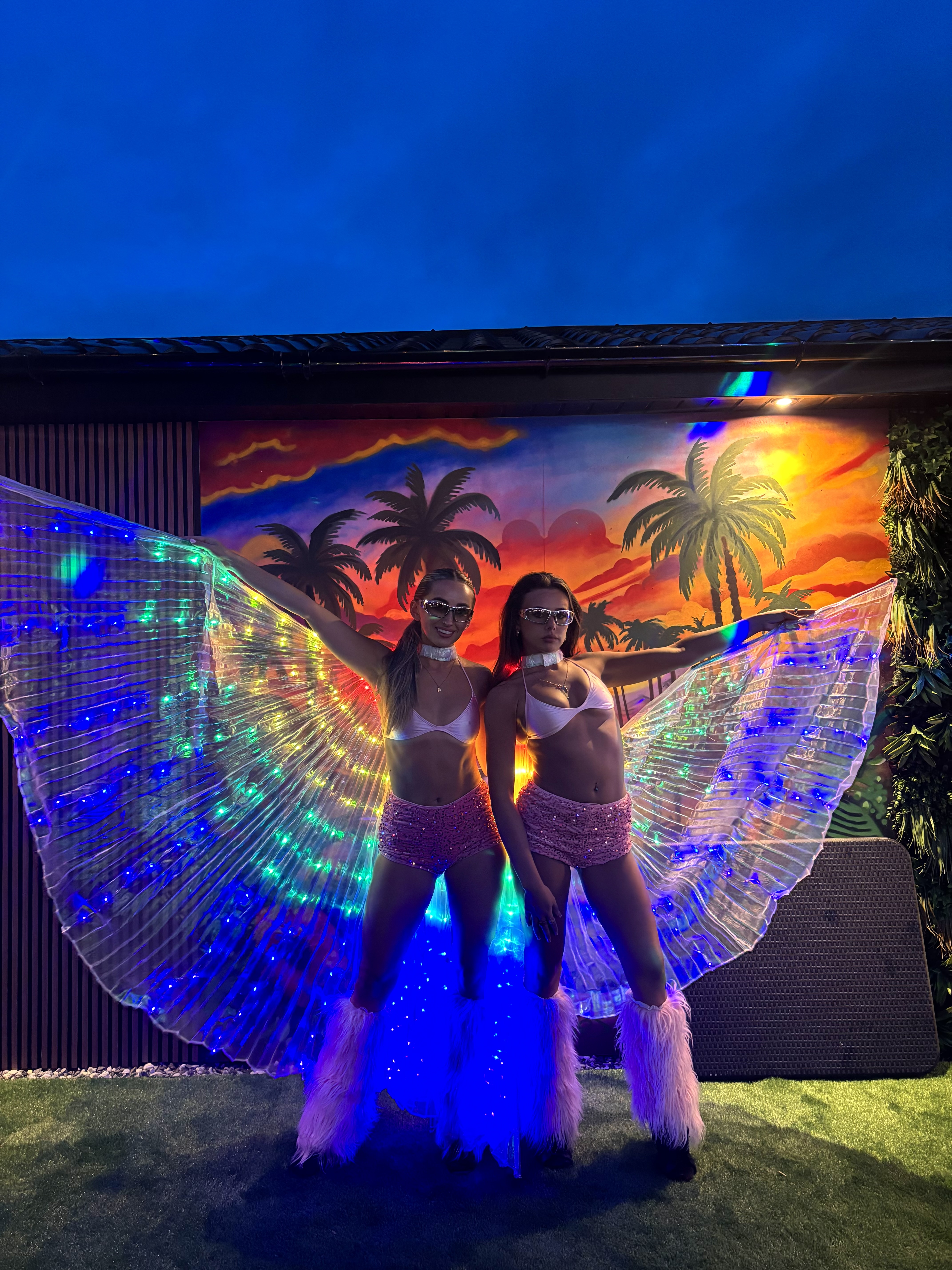 Two dancers pose with rainbow LED wings before a tropical sunset mural, pink sequins, and moody blue event lighting.