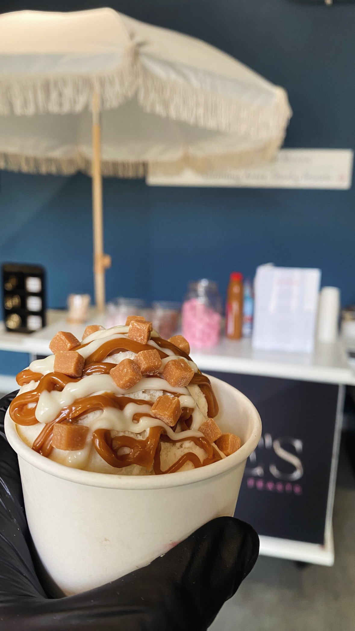 A gloved hand holds a cup of vibrant ice cream topped with red drizzle and white chocolate, set against a sleek, modern serving station.