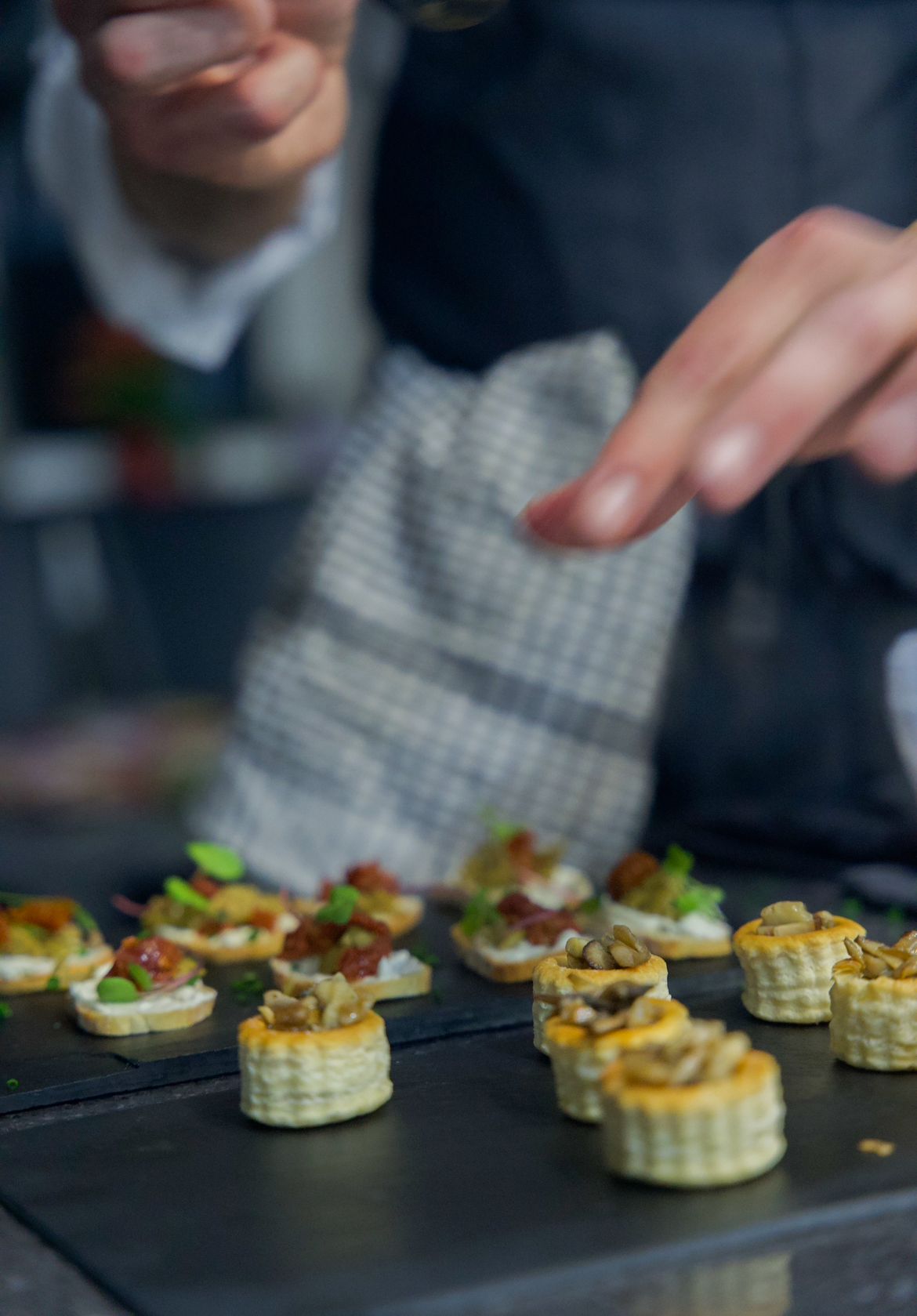 Delicate canapés, topped with herbs and toppings, are artfully arranged on a dark slate platter with warm evening lighting enhancing their appeal.