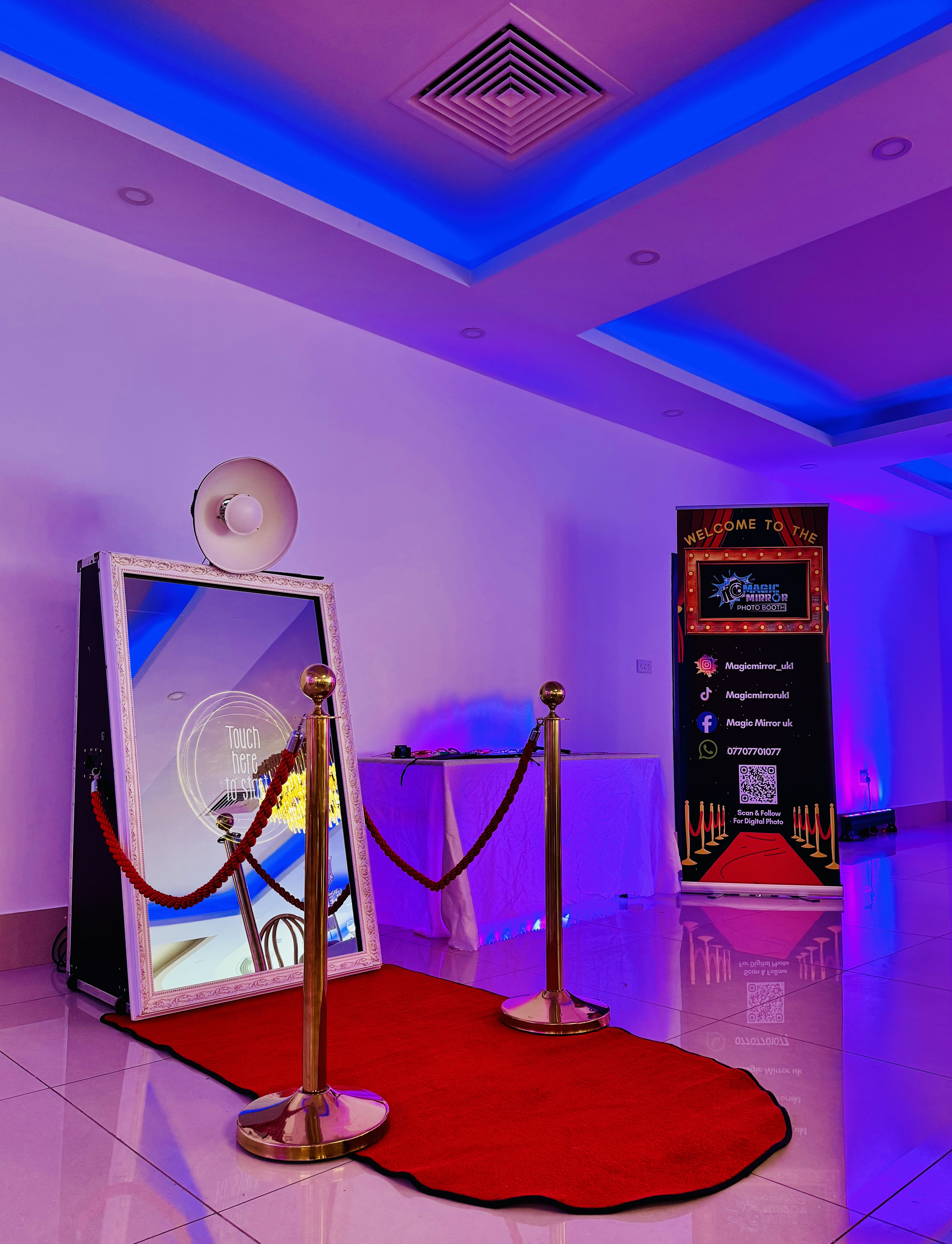 A glam event setup features a red carpet leading to a mirror photo booth, framed elegantly, under vibrant purple LED lighting.