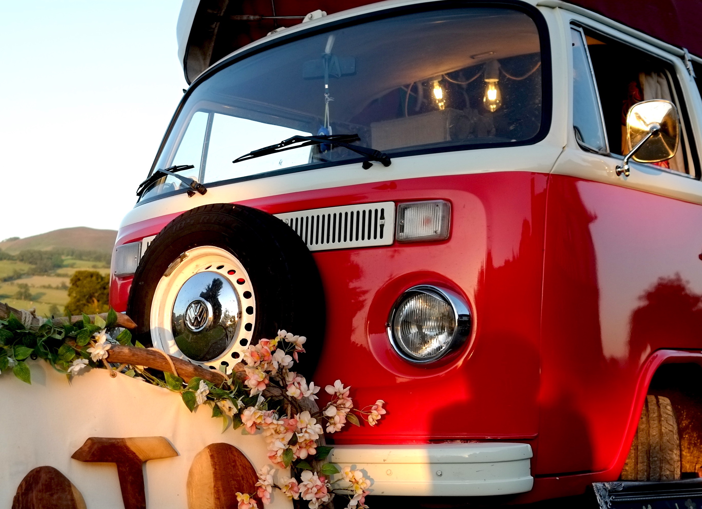 A vintage red VW van adorned with pink flowers and rustic wood accents, illuminated by warm evening light, evokes a charming outdoor catering vibe.