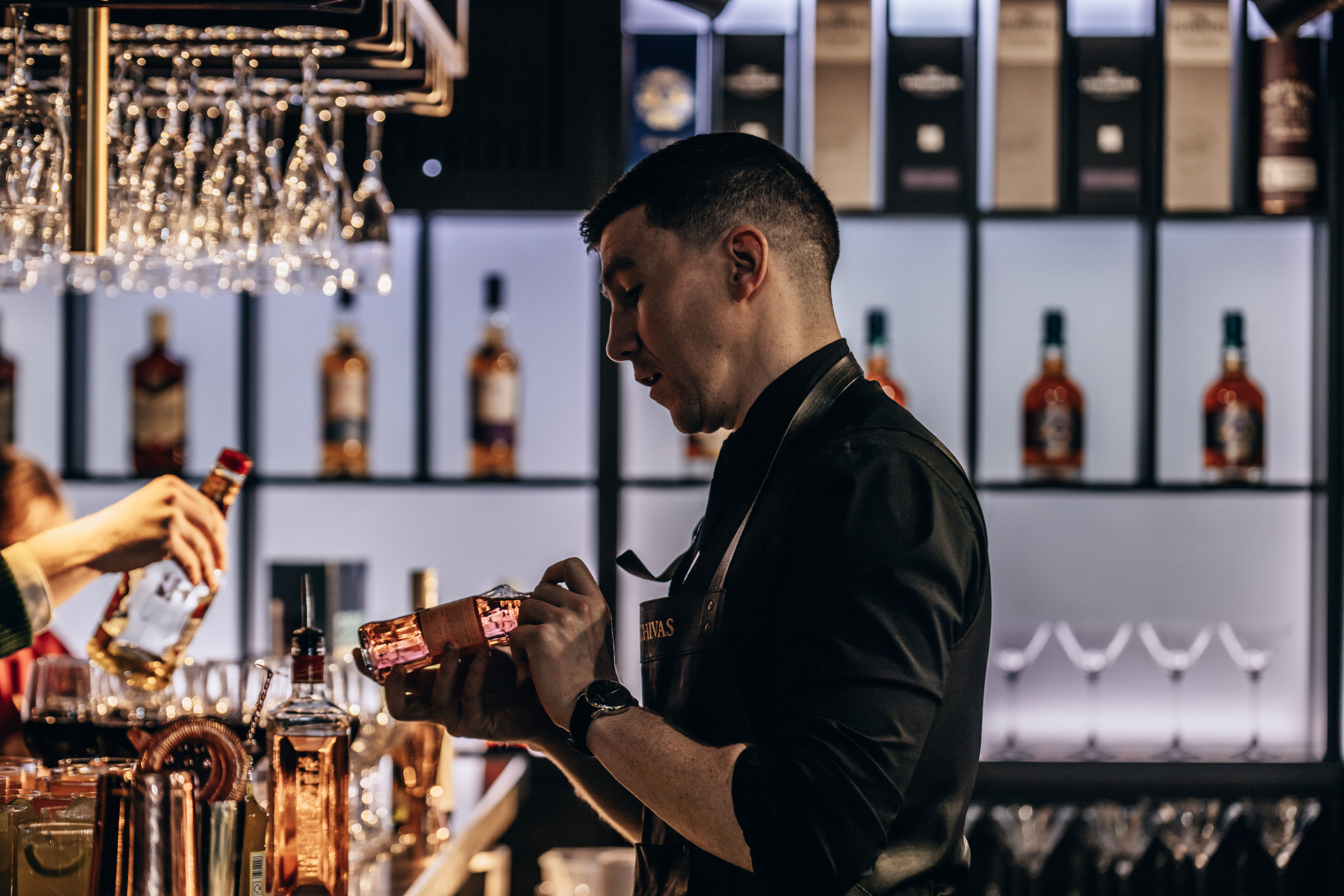 A cheerful bartender stands behind a rustic wooden bar, showcasing spirits and cocktail tools under bright daylight, enhancing the inviting atmosphere.