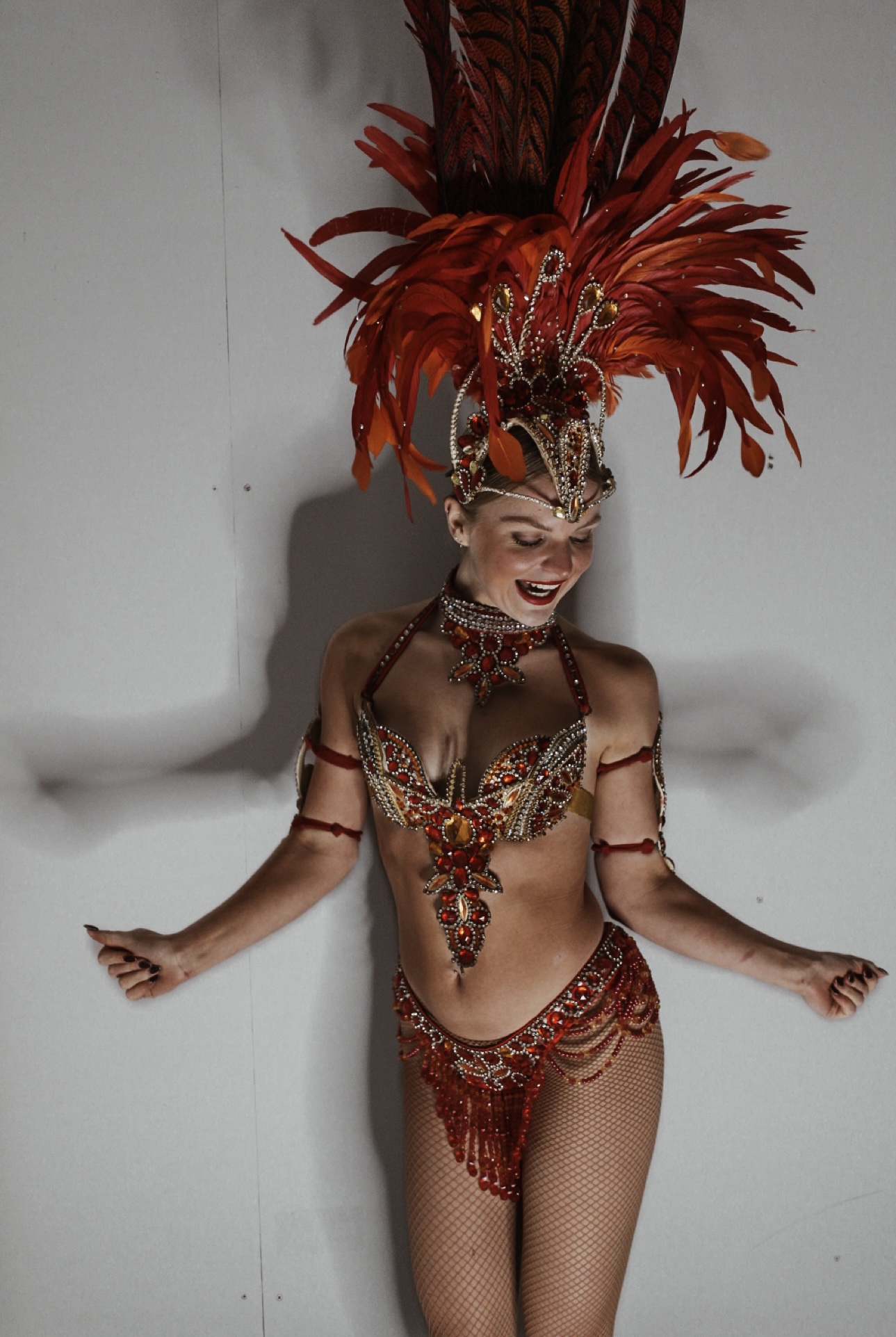 A performer in a dazzling red and gold costume with intricate beading and feathers exudes vibrant energy against a neutral backdrop.