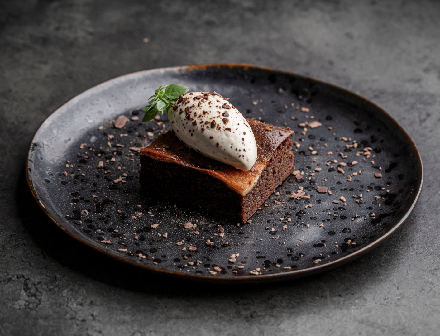 A rich chocolate brownie is elegantly plated on a textured black ceramic dish, topped with creamy vanilla ice cream and chocolate shavings, under soft, moody lighting.