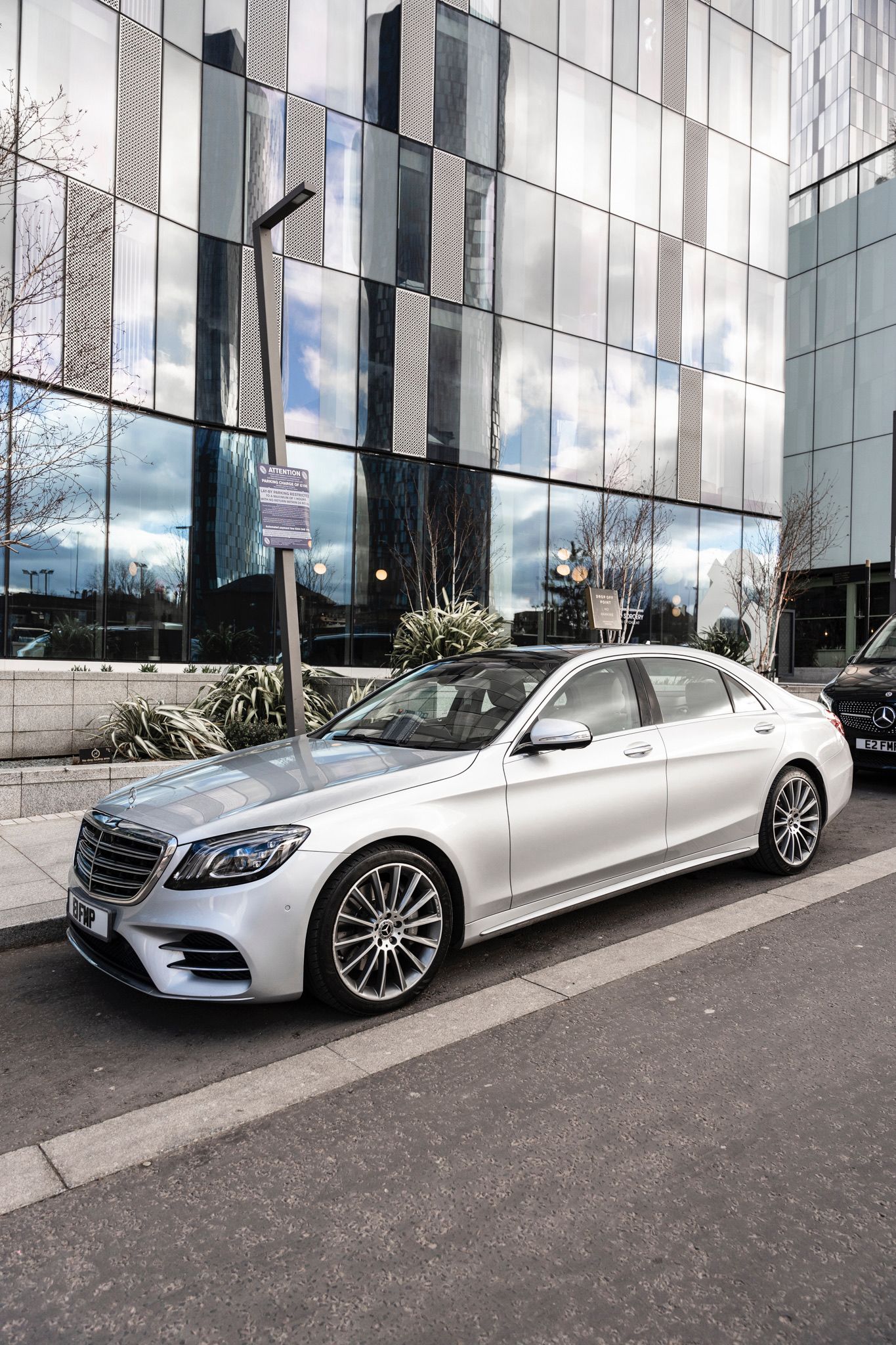 A sleek silver Mercedes sedan rests on a modern urban street, reflecting glass facades and surrounded by elegant greenery and bright daylight.