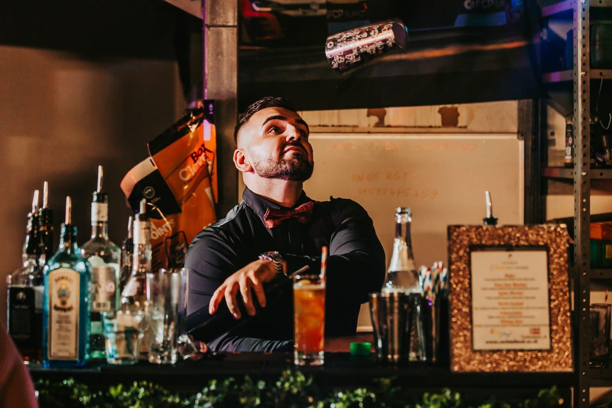 A skilled bartender, dressed in a black shirt and bow tie, juggles a cocktail shaker against a cozy, softly lit backdrop of colorful bottles.
