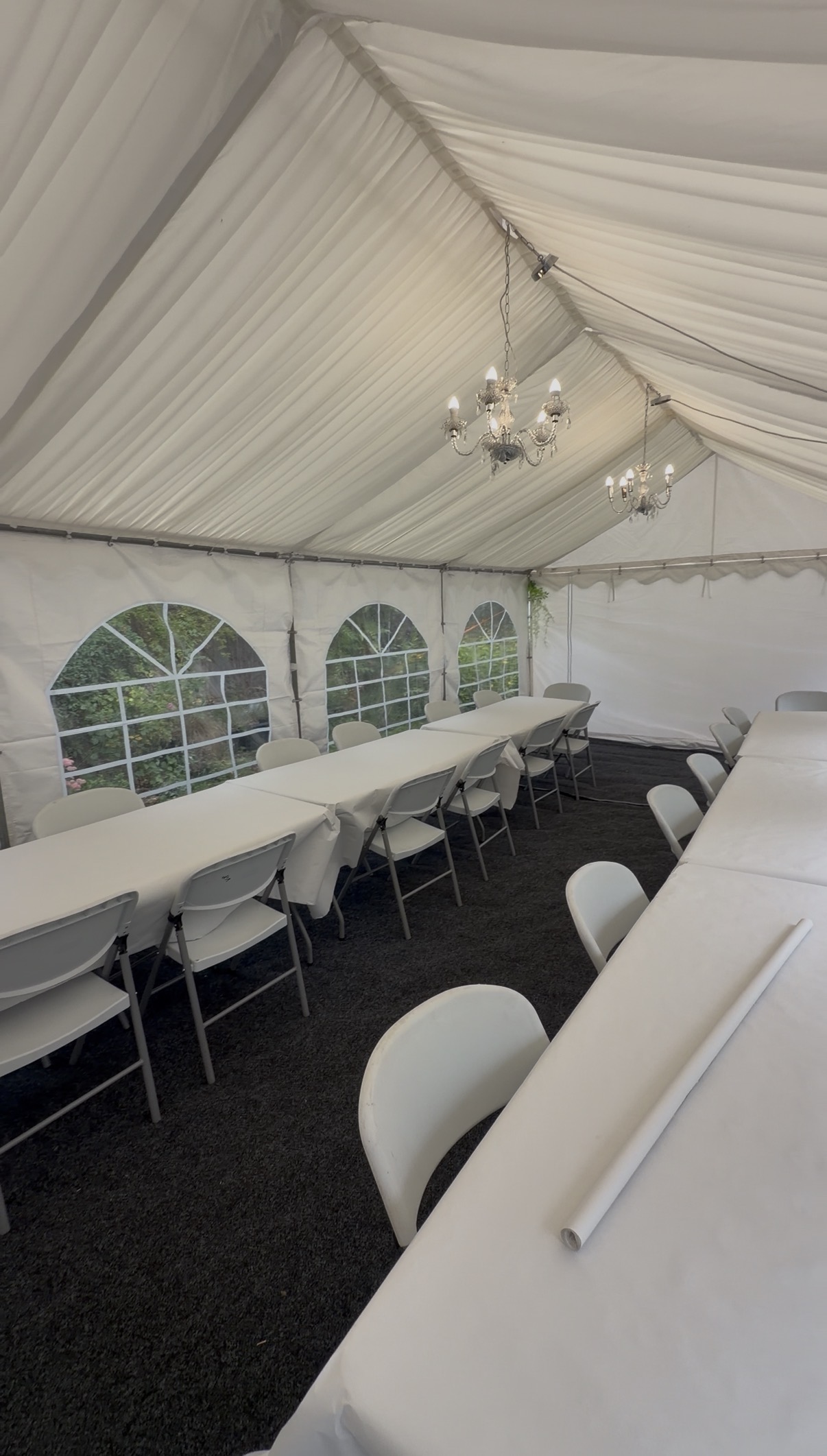 Elegant wedding catering tent with white draped ceiling, chandelier lights, banquet tables, and folding chairs on dark carpet in soft daylight.