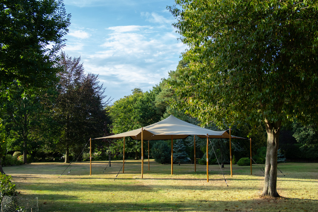 Elegant beige stretch tent on a manicured lawn, framed by lush trees and blue sky, ideal for upscale outdoor catering service.