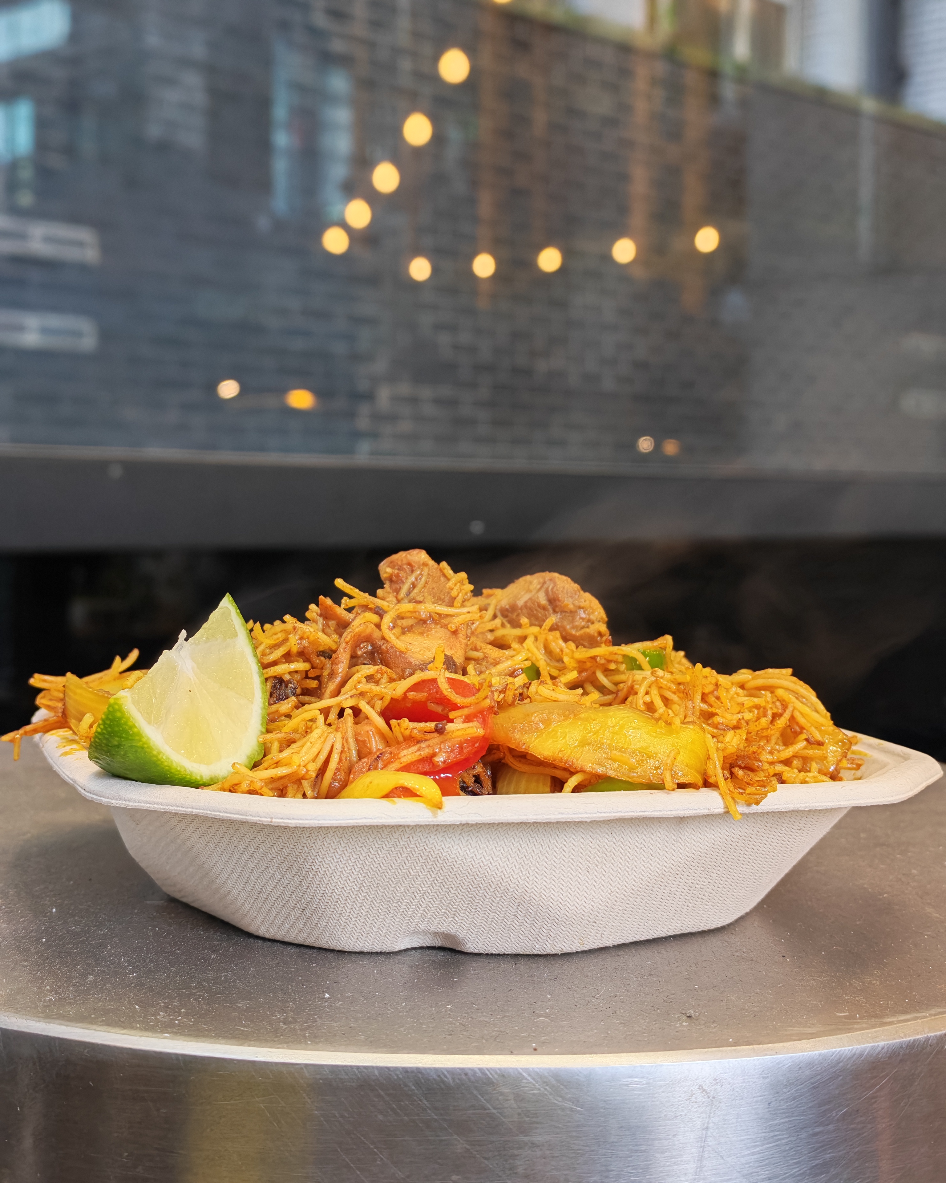 A warm, savory dish of stir-fried noodles with chicken, vibrant bell peppers, and a fresh lime wedge, served on eco-friendly paperware.