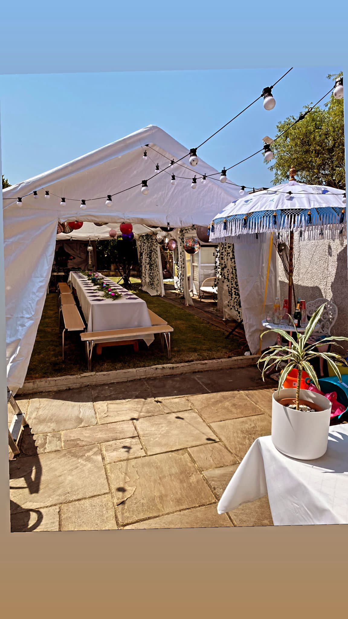 A bright outdoor catering setup features a long, white table adorned with colorful flowers, under a tent with decorative string lights, creating a festive atmosphere.