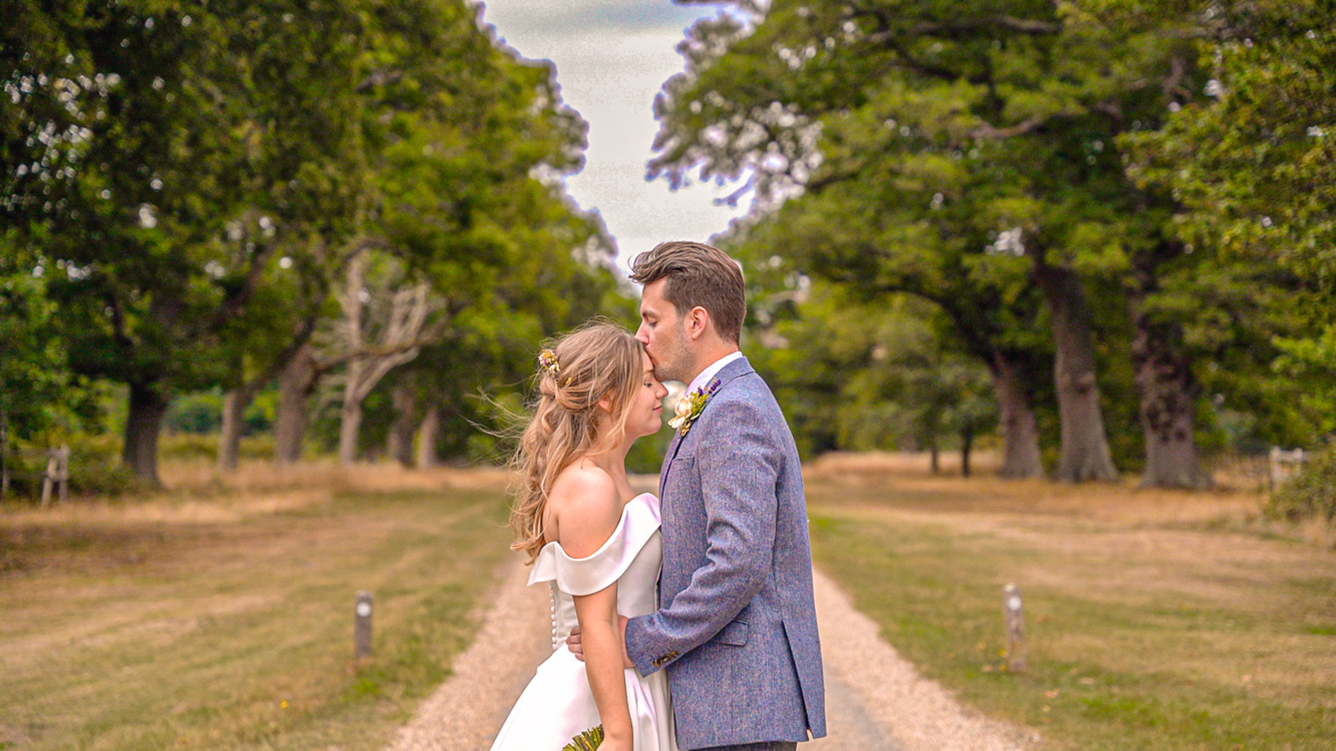 Romantic outdoor wedding portrait: a groom in blue tweed kisses a bride in an off-shoulder white gown on a tree-lined gravel path, soft daylight.