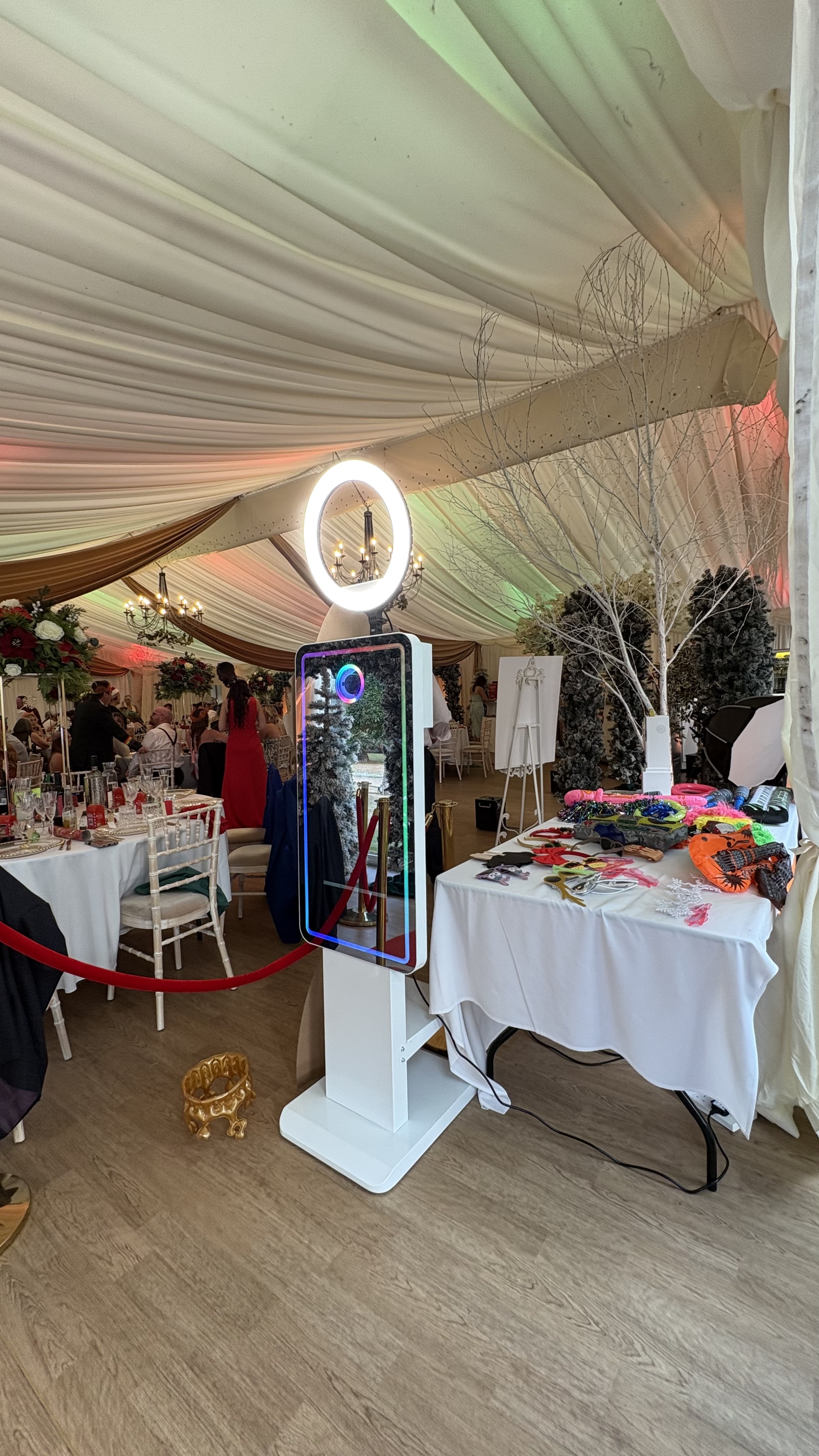 Elegant wedding photo booth in a draped tent, with LED ring light, white tables, props, and warm chandeliers for lively catering.