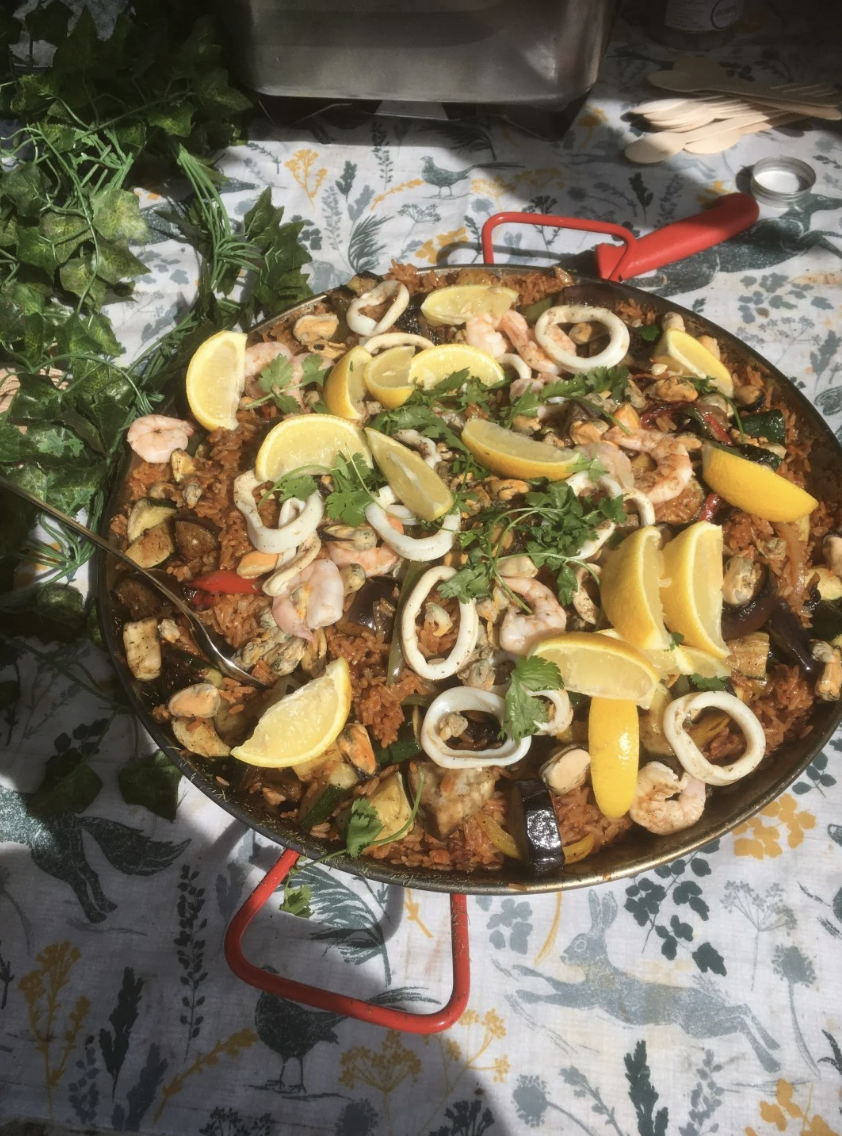 Sunlit catering paella in a black pan with red handles, shrimp, squid, lemon wedges, and parsley on a patterned linen buffet table.