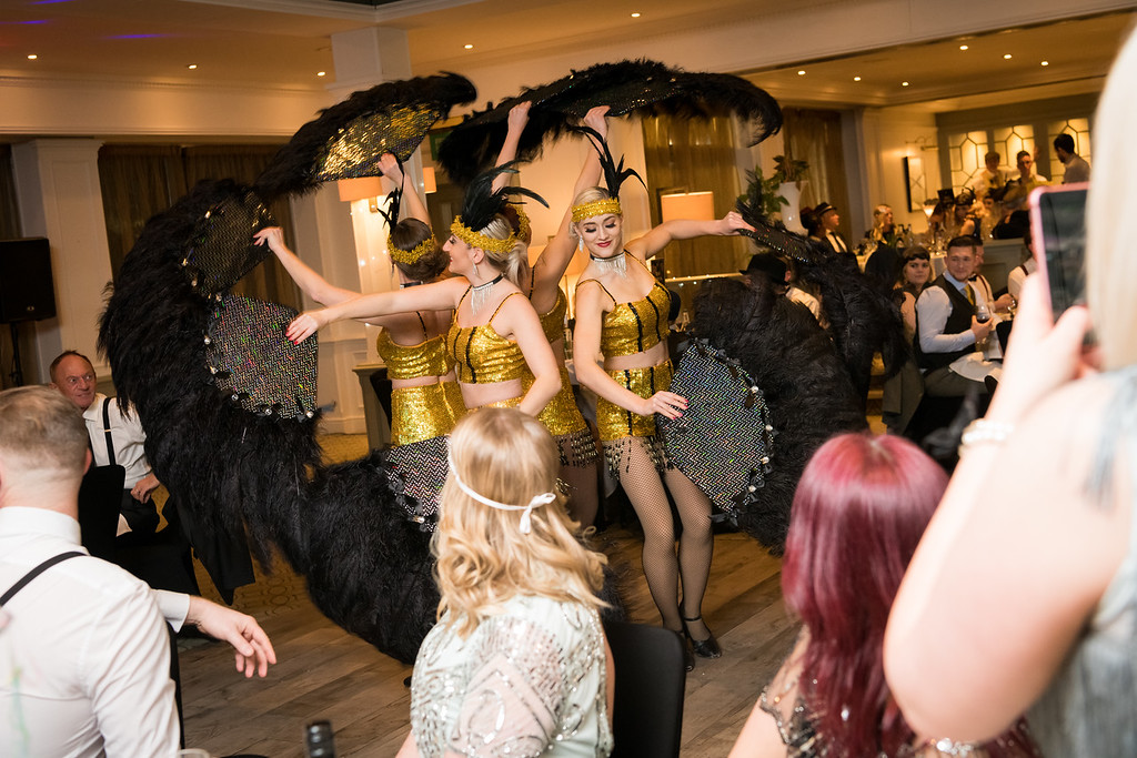 Glistening in gold sequins, dancers showcase vibrant feather fans amid softly lit, elegant surroundings, energizing the upscale event ambiance.