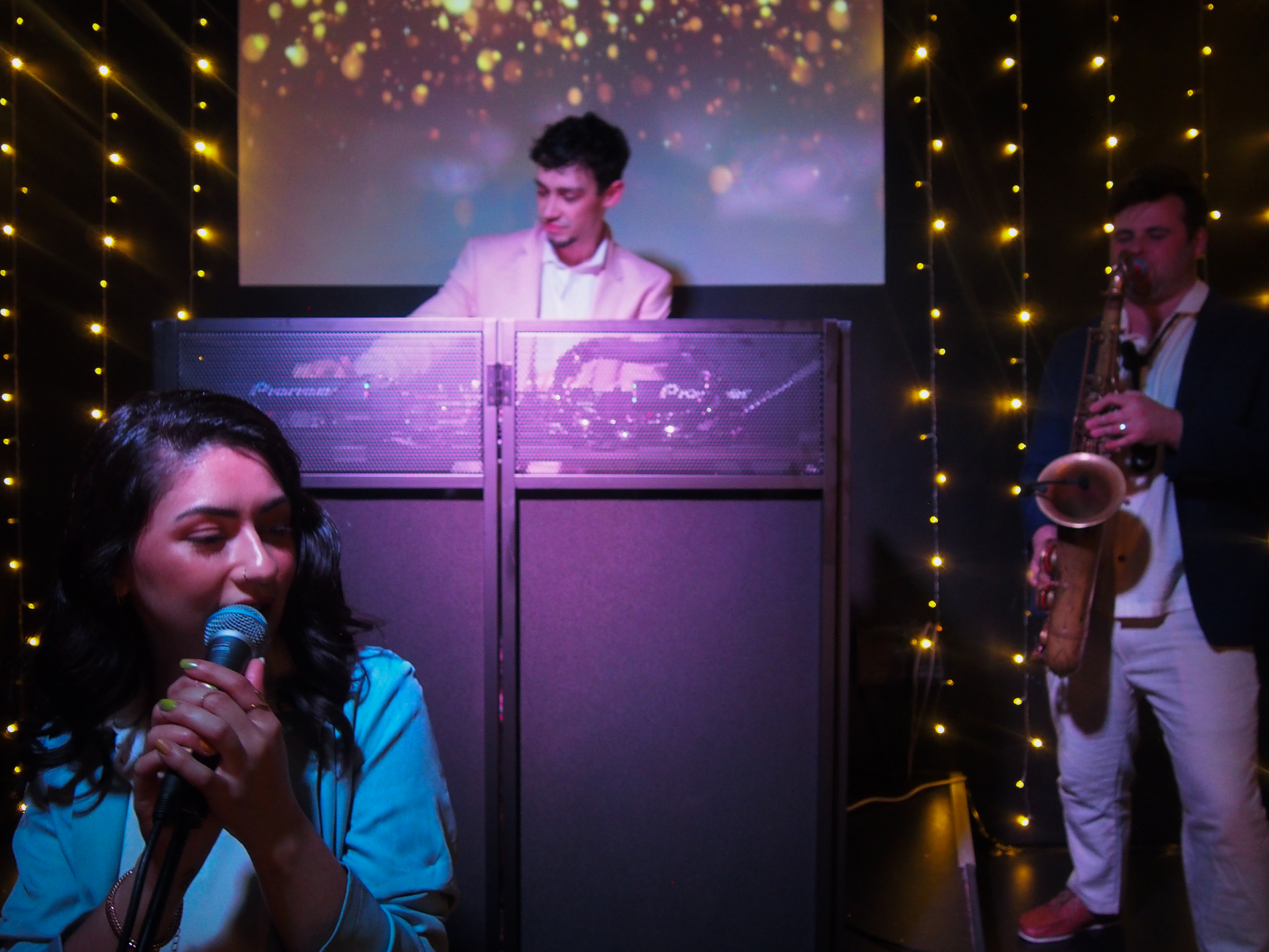 A vibrant performance scene features a singer with a microphone, a DJ in a stylish suit, and a saxophonist, all amid sparkling string lights.