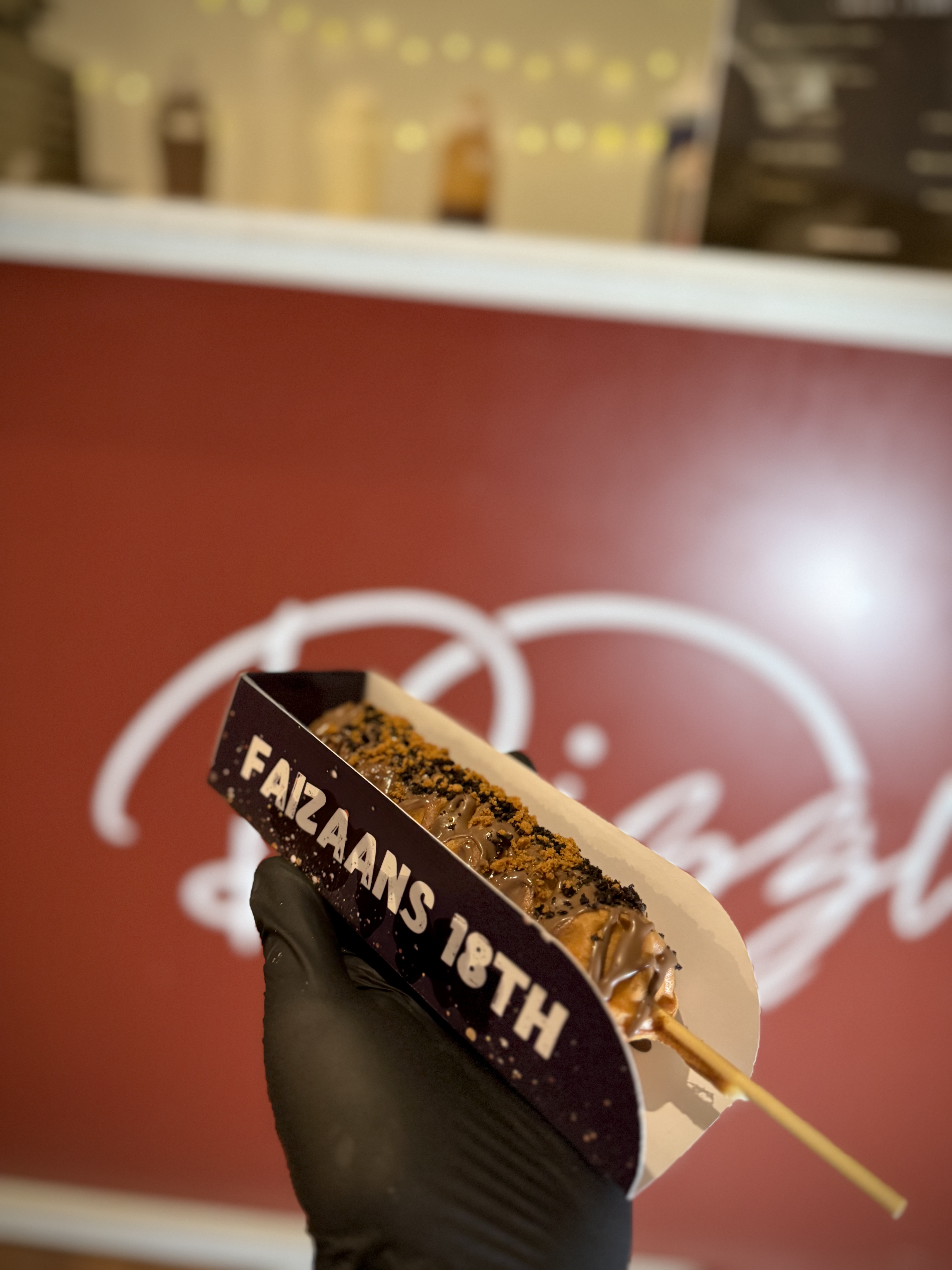A chocolate-covered treat with crushed cookies sits in a branded box, held by a gloved hand; a vibrant red backdrop adds festive flair.