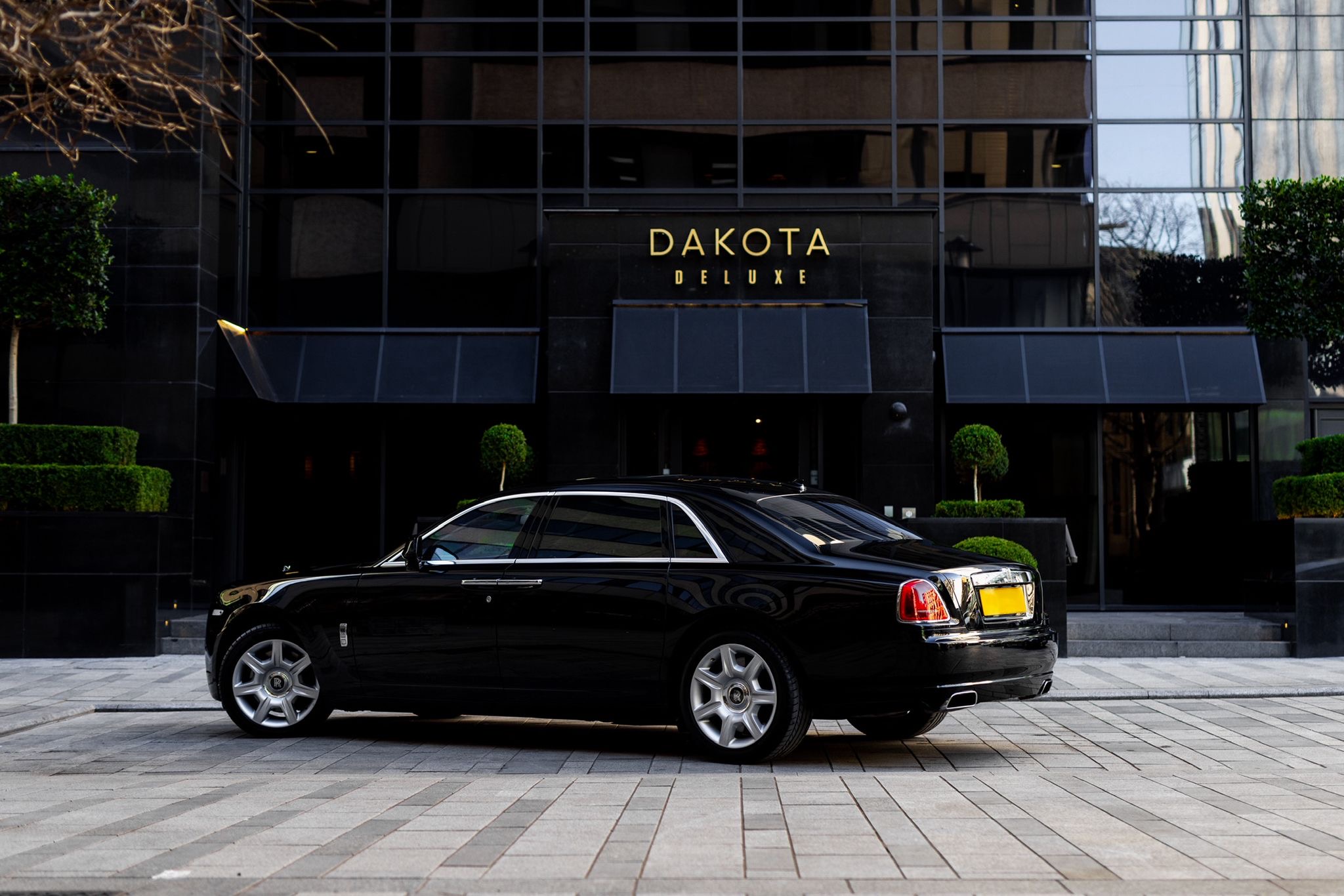 A sleek black luxury car is parked in front of the upscale Dakota Deluxe hotel, highlighted by polished stone pavers and manicured shrubs, exuding elegance.