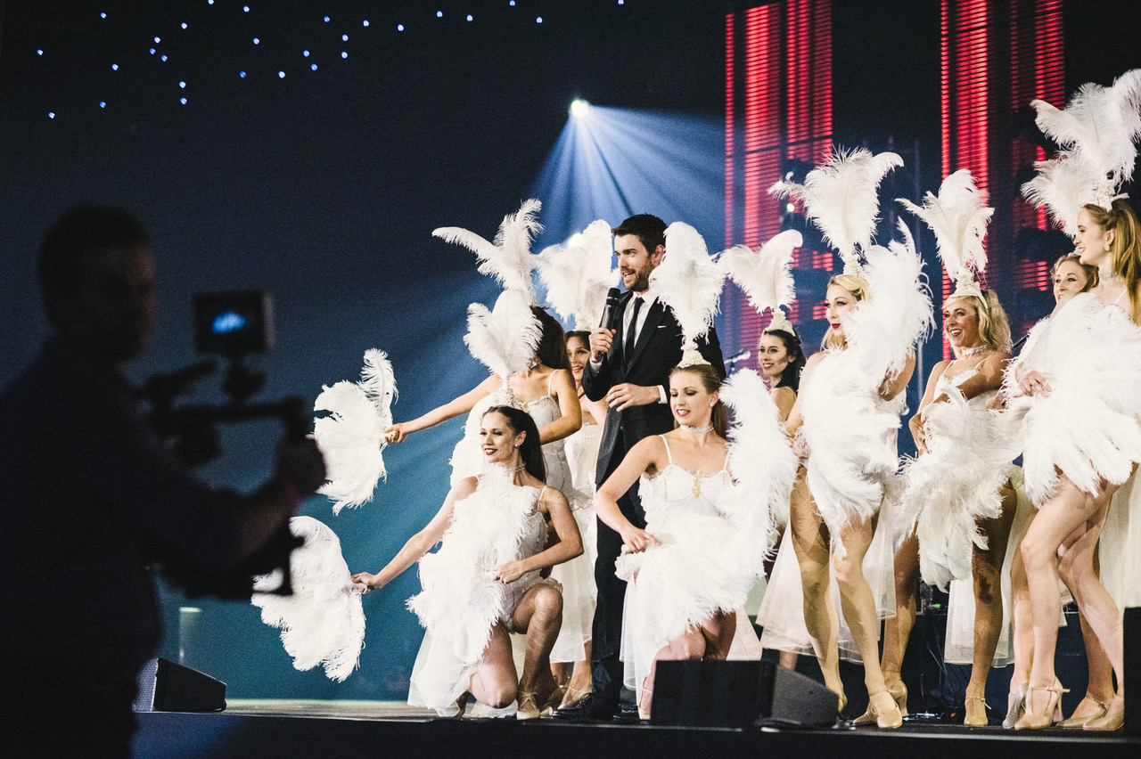 A dynamic stage scene features a sharply dressed host and dancers in elegant white costumes, with feather boas and radiant smiles under vibrant lights.