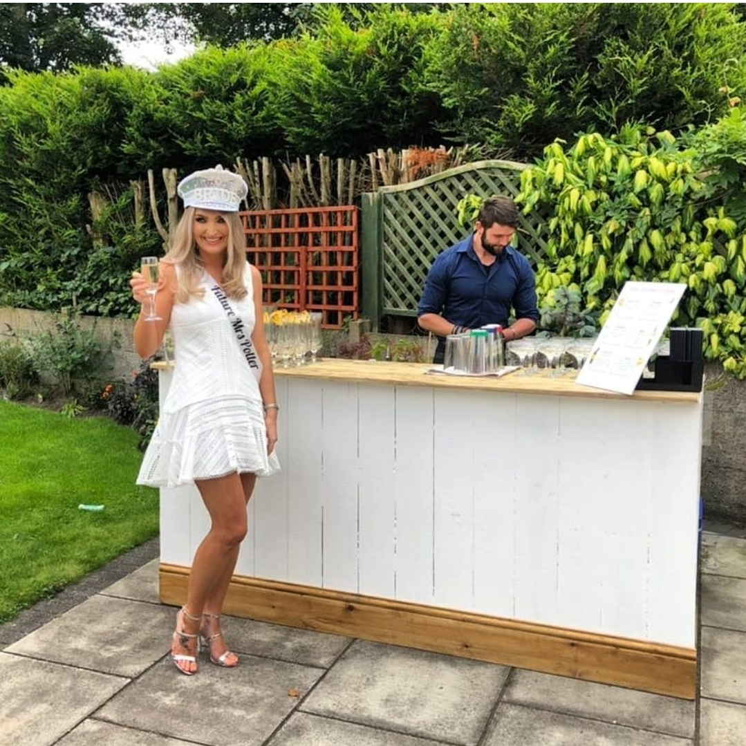 A stylish outdoor bar features a smiling woman in a white dress and sash, enjoying a drink as a bartender prepares cocktails beside her.