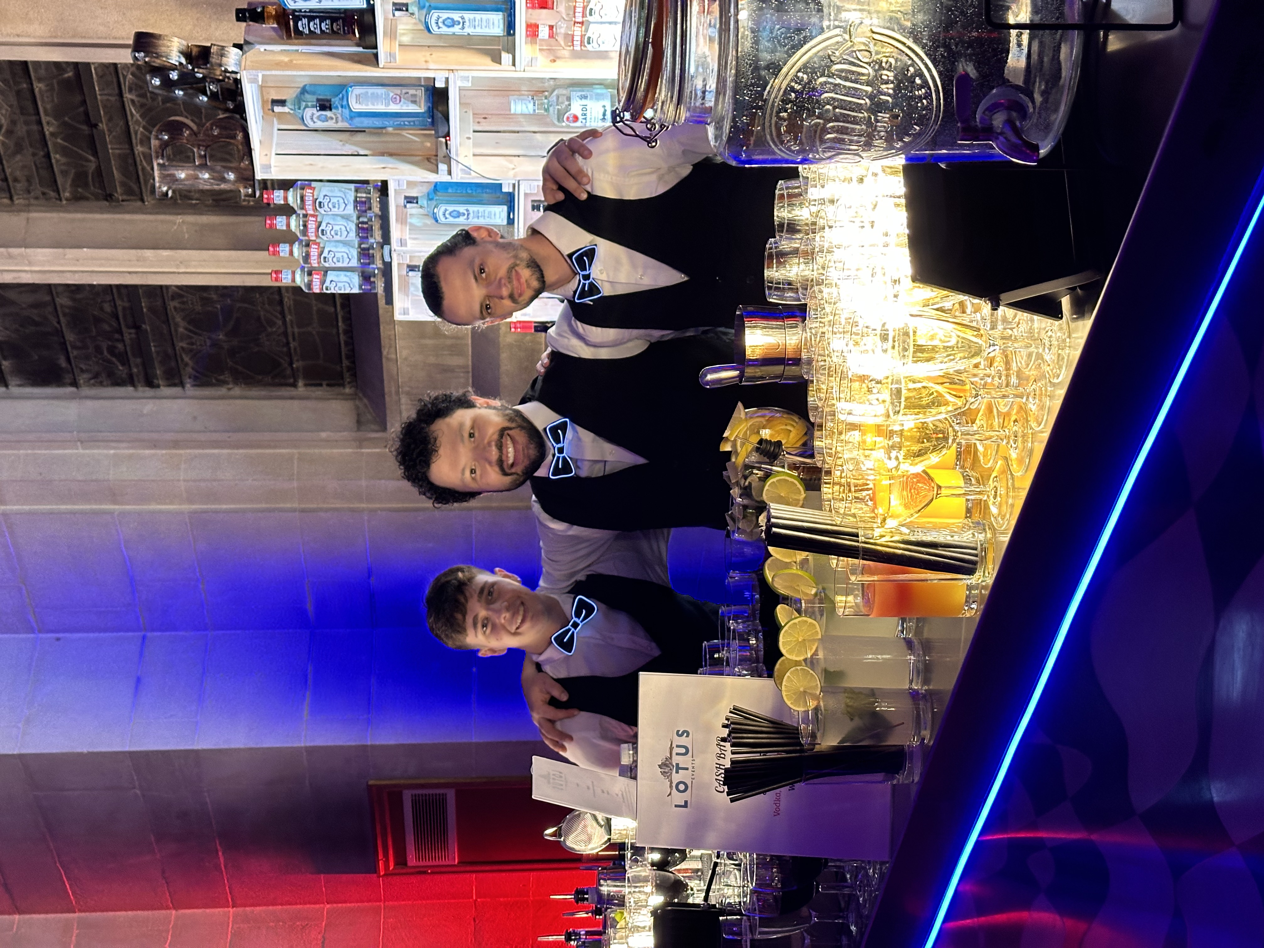 Three bartenders in sleek black vests and bowties stand at a vibrant, illuminated bar with colorful drinks, enhancing the lively atmosphere.