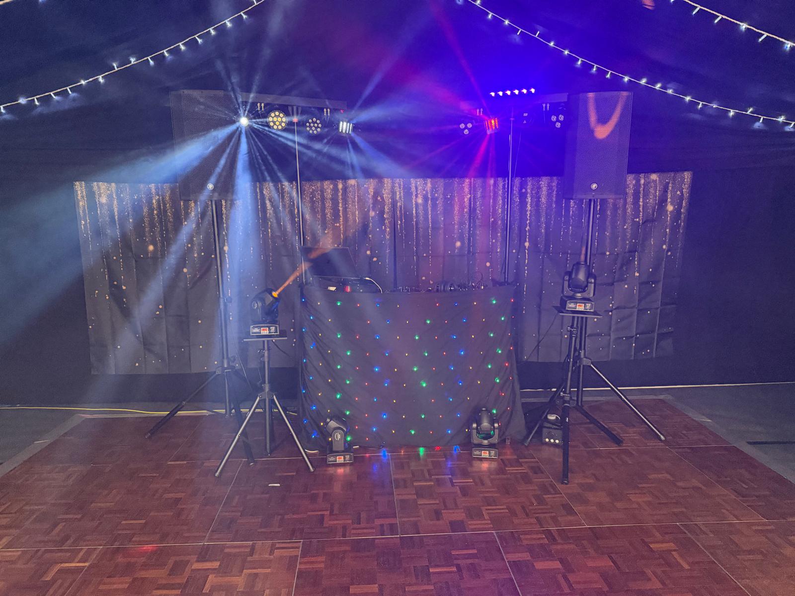 Professional DJ booth with LED-lit black drape, twin speakers, and moving lights on a parquet dance floor under moody string lights.