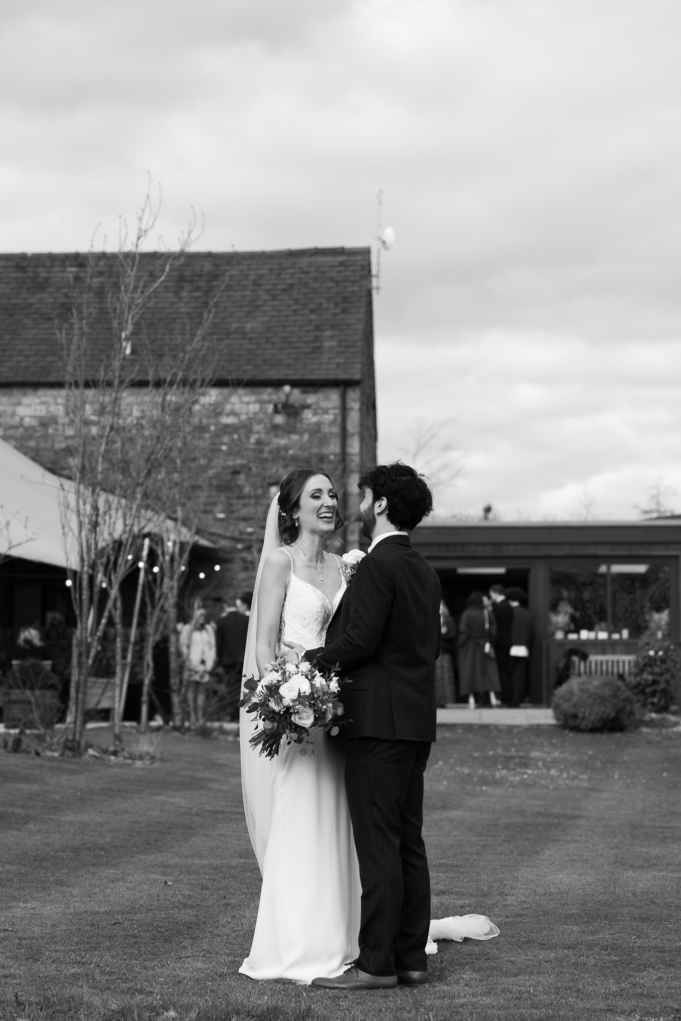 A smiling couple, dressed elegantly, share an intimate moment outdoors, surrounded by rustic architecture and joyful guests, capturing wedding bliss.