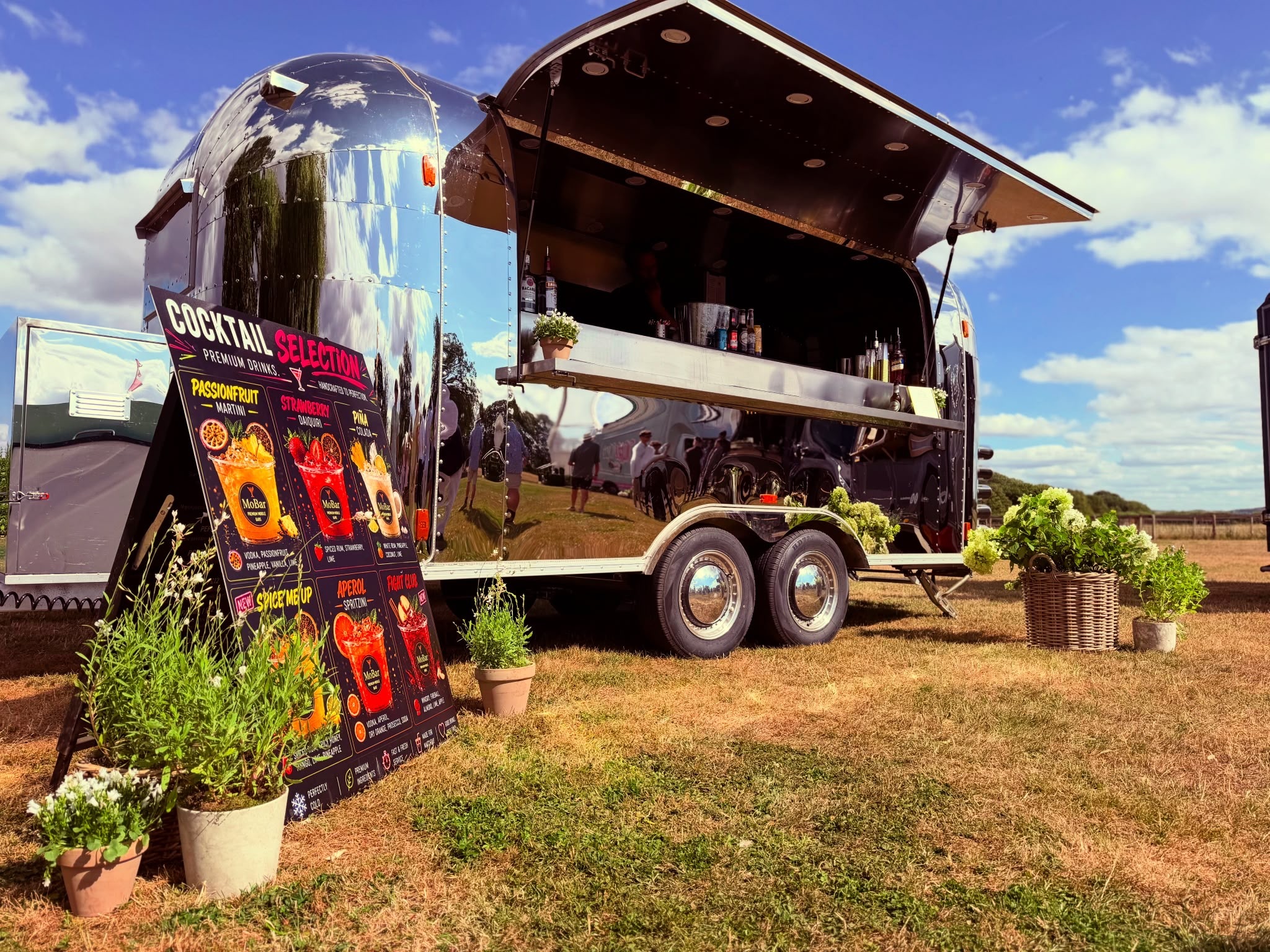 Polished vintage cocktail trailer with chrome finish, open serving hatch, stacked cups and drink bottles, on sunny lawn.