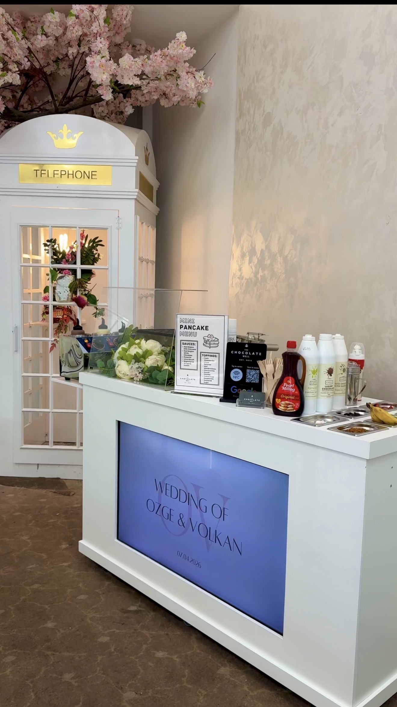 Elegant wedding pancake catering station with a white London-phone-booth backdrop, pink cherry blossoms, and neatly arranged toppings in warm indoor light.