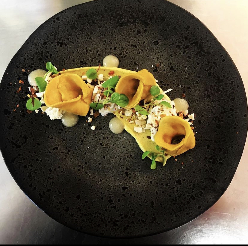 Artfully presented on a textured black plate, the dish features yellow dumplings, creamy cheese, microgreens, and delicate sauce drops, creating an elegant, refined dining experience.