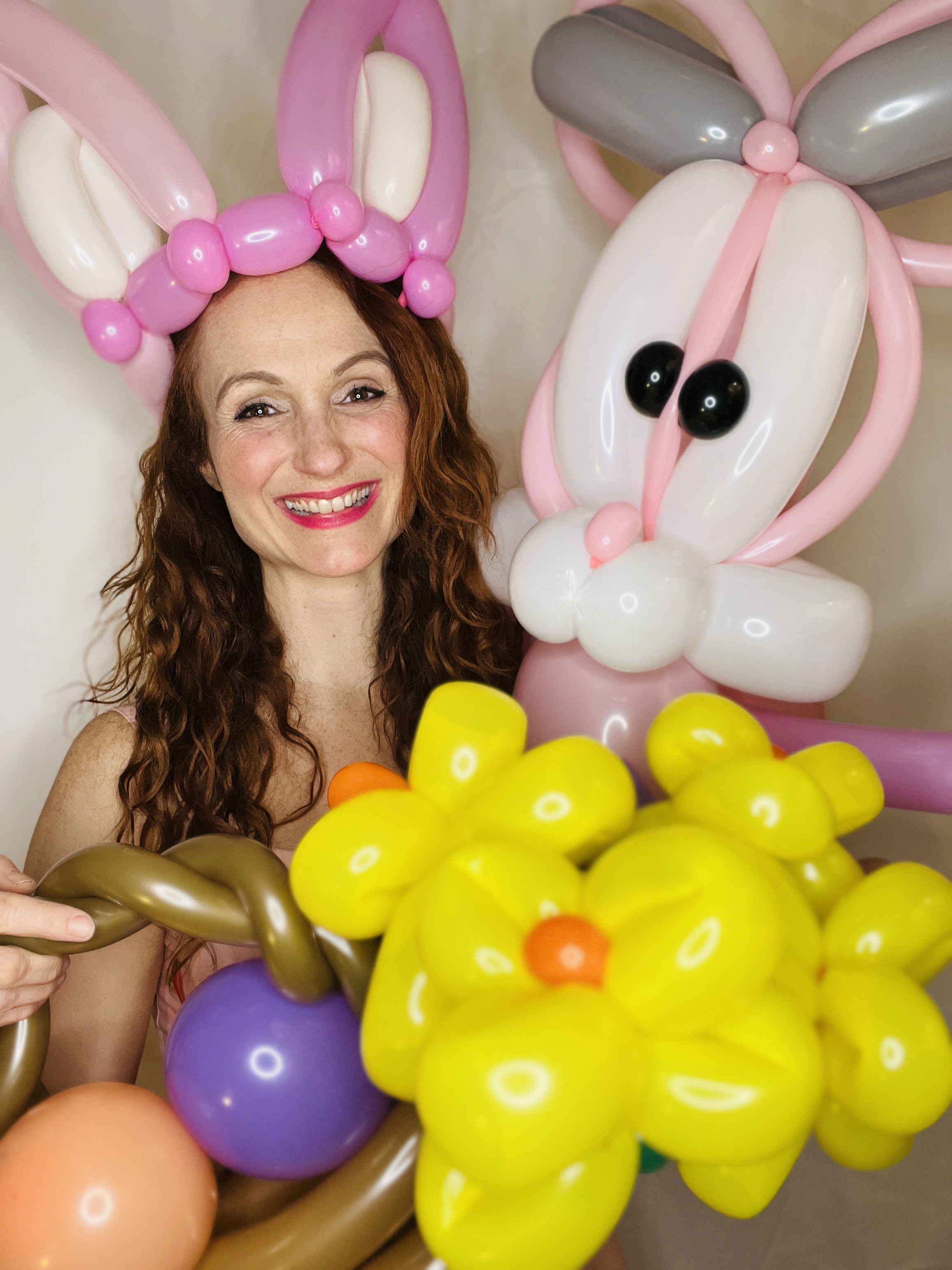 A smiling woman wears a pink and white balloon bunny hat, holding a vibrant bouquet of yellow, purple, and orange balloon flowers, exuding joy.