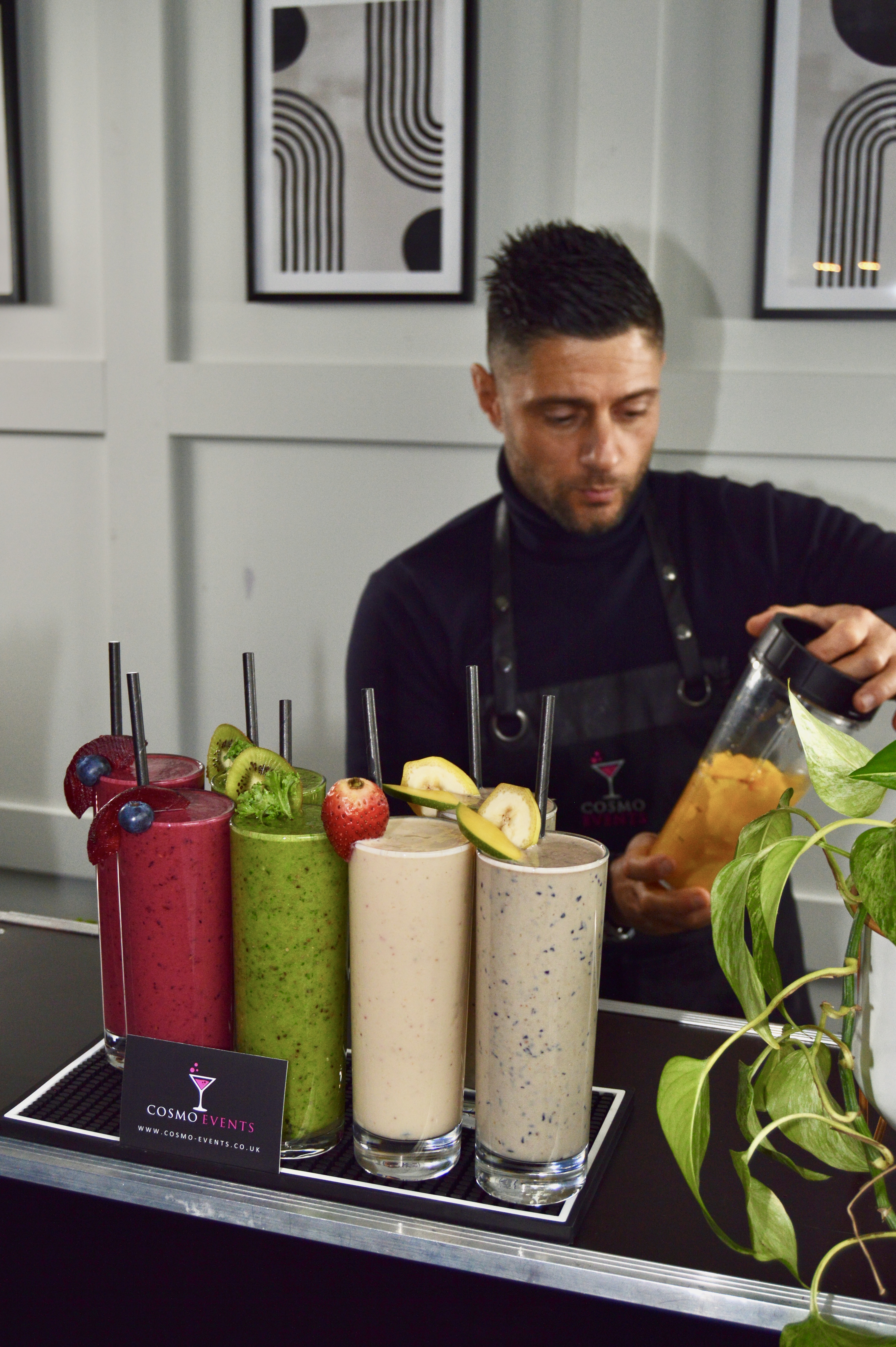 A bartender prepares vibrant smoothies in glass containers, featuring colors like deep red, green, and beige, against a sleek black backdrop.