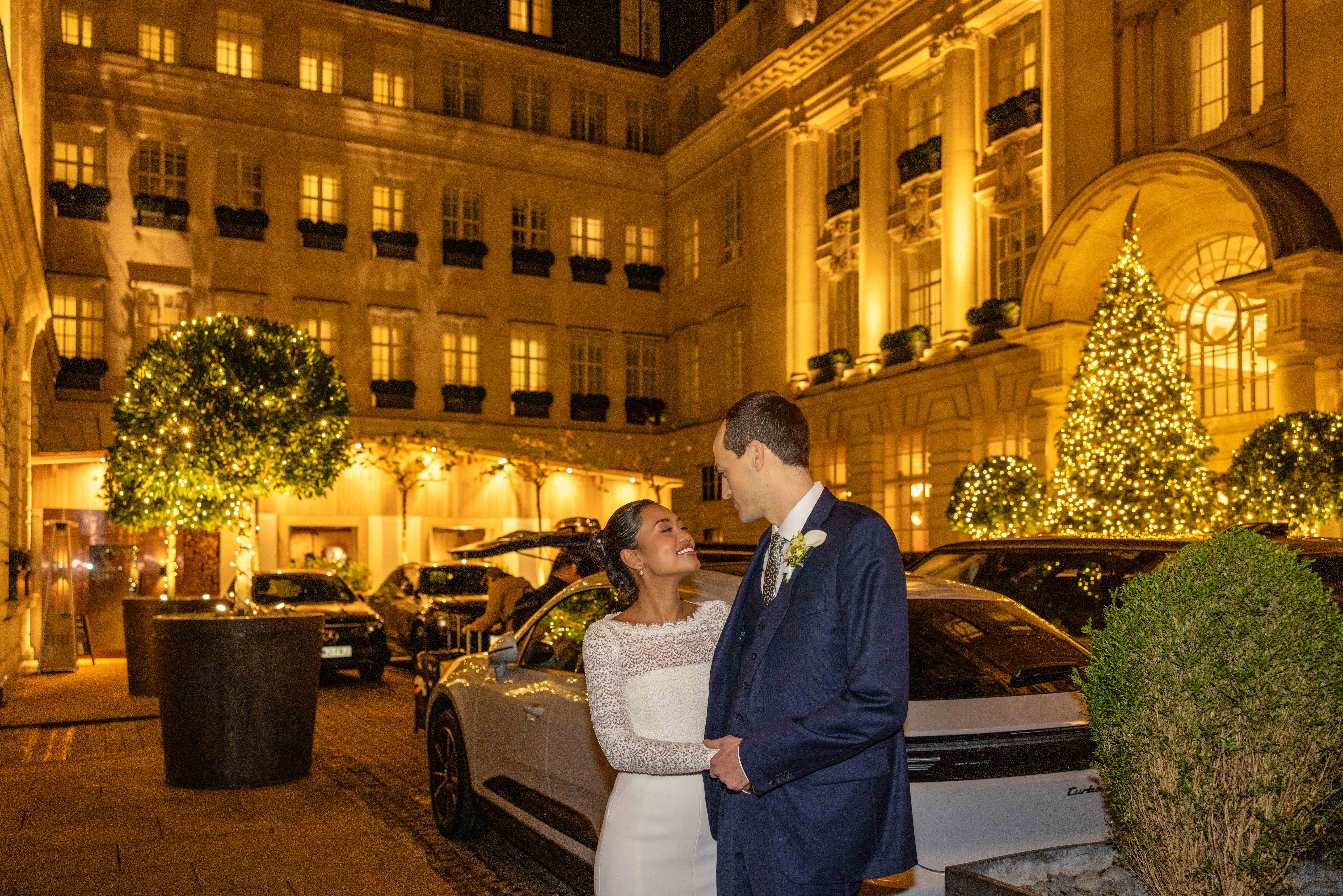 A beautifully lit hotel courtyard features a happy couple dressed elegantly, surrounded by lush greenery and softly glowing Christmas lights, enhancing the romantic atmosphere.
