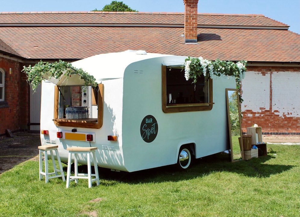 A vintage white bar trailer with wooden accents sits on lush green grass, featuring floral garlands above its open window, radiating a cheerful, outdoor catering vibe.