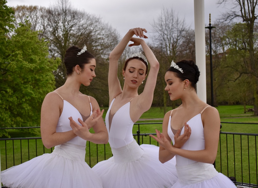 Three graceful ballerinas in white tutus pose elegantly outdoors, surrounded by lush greenery, exuding a serene, artistic atmosphere.