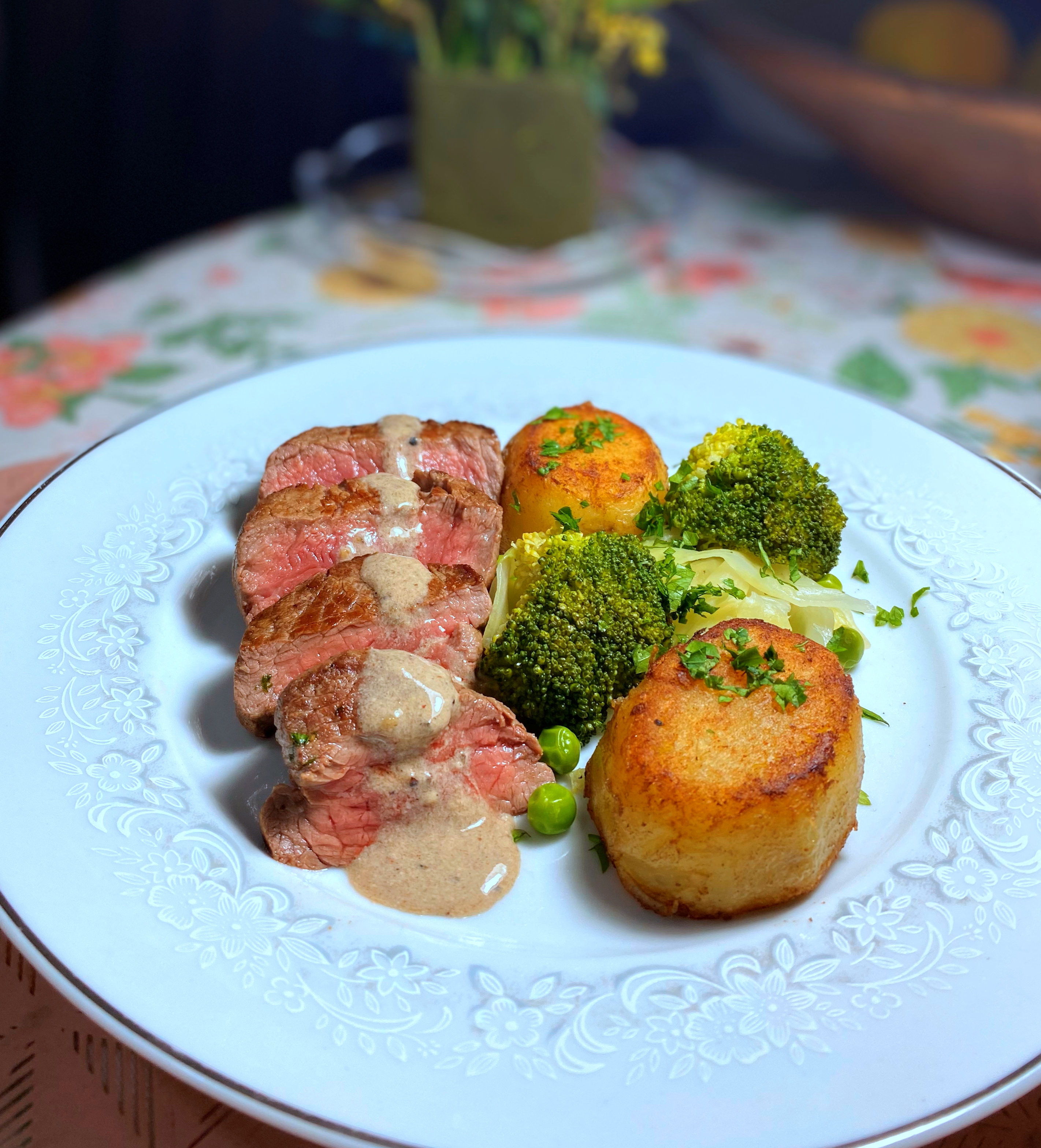 A beautifully arranged plate features tender beef slices drizzled with creamy sauce, surrounded by golden roasted potatoes and vibrant green broccoli, set on a delicate floral plate.