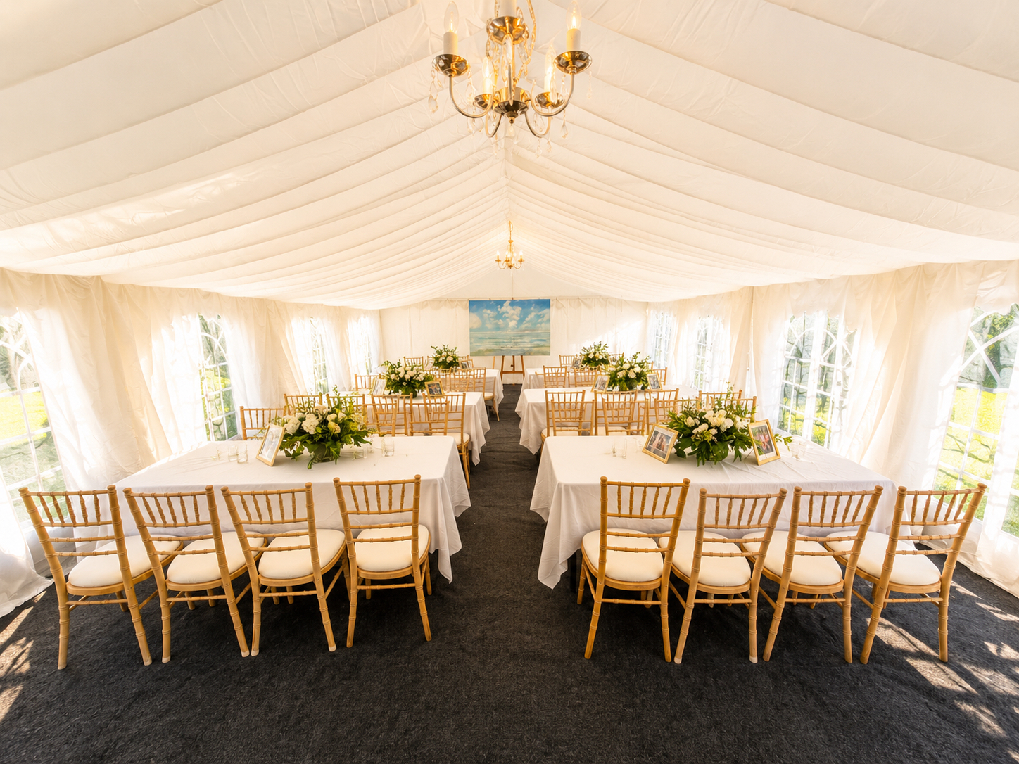 Bright daylight floods a white draped marquee with gold Chiavari chairs, linen tables, floral centerpieces, and elegant catering setup.