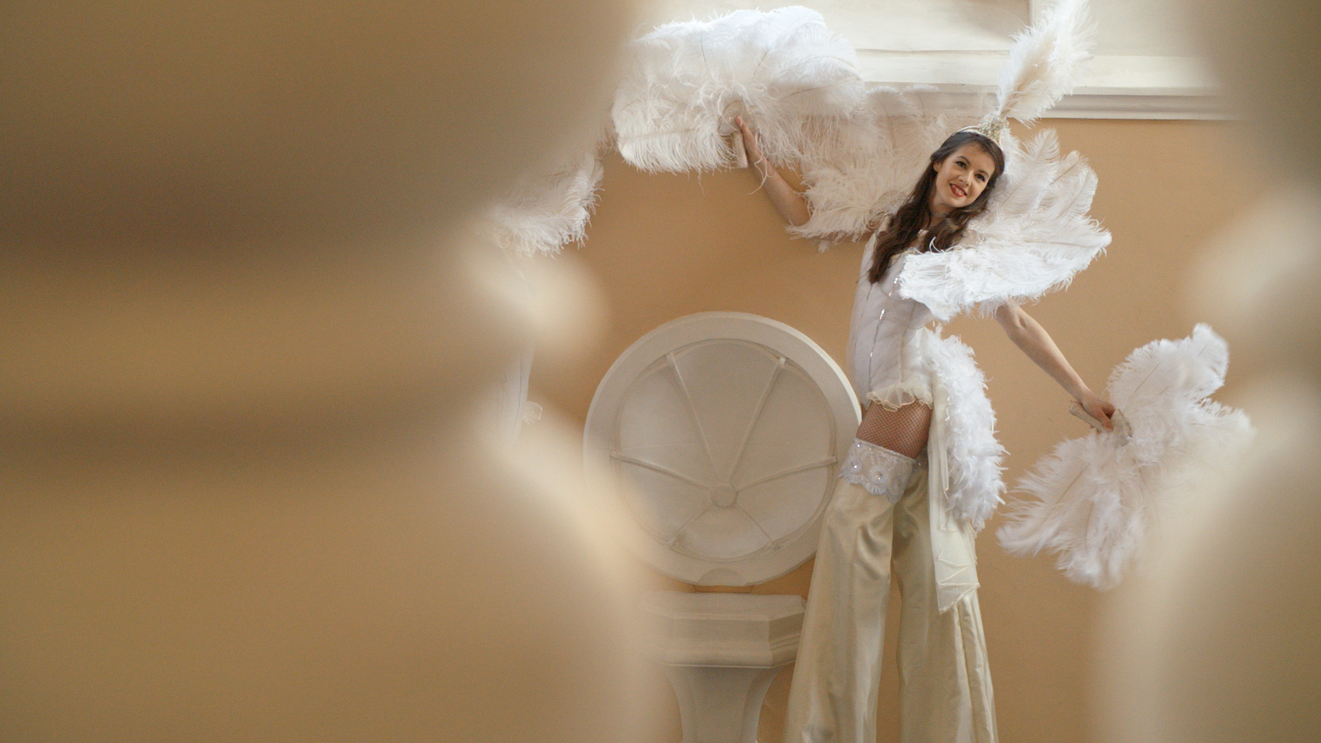 A performer in elegant white feather attire playfully poses against a warm, peach-colored wall, creating a vibrant, celebratory atmosphere.