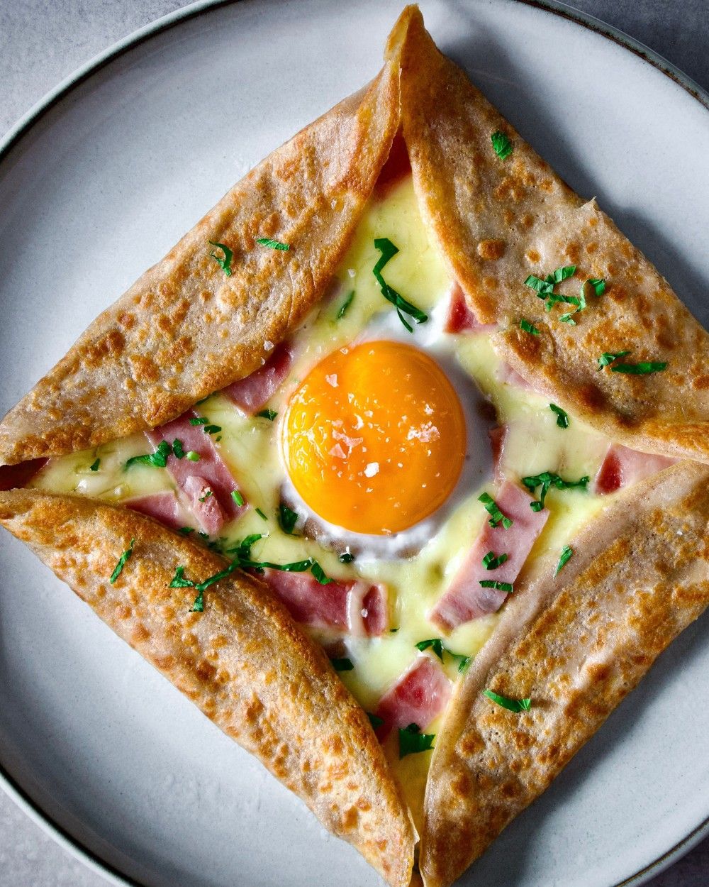 A beautifully crafted buckwheat galette features melted cheese, ham, vibrant herbs, and a perfectly runny egg yolk, presented artfully.
