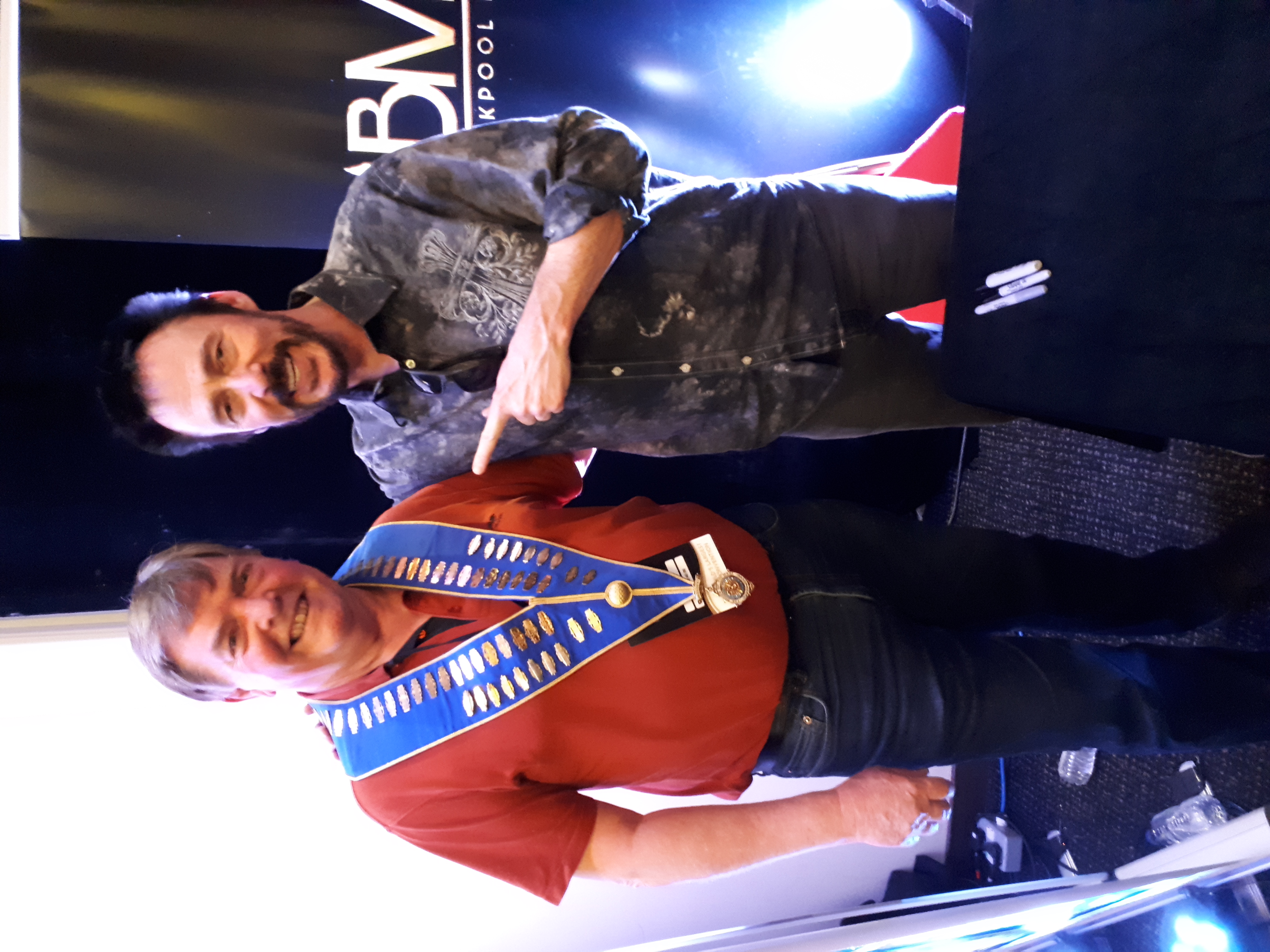 Two men smile widely in a casual indoor setting, one wearing a decorated blue sash, with a backdrop of dim lighting and softly glowing colors.